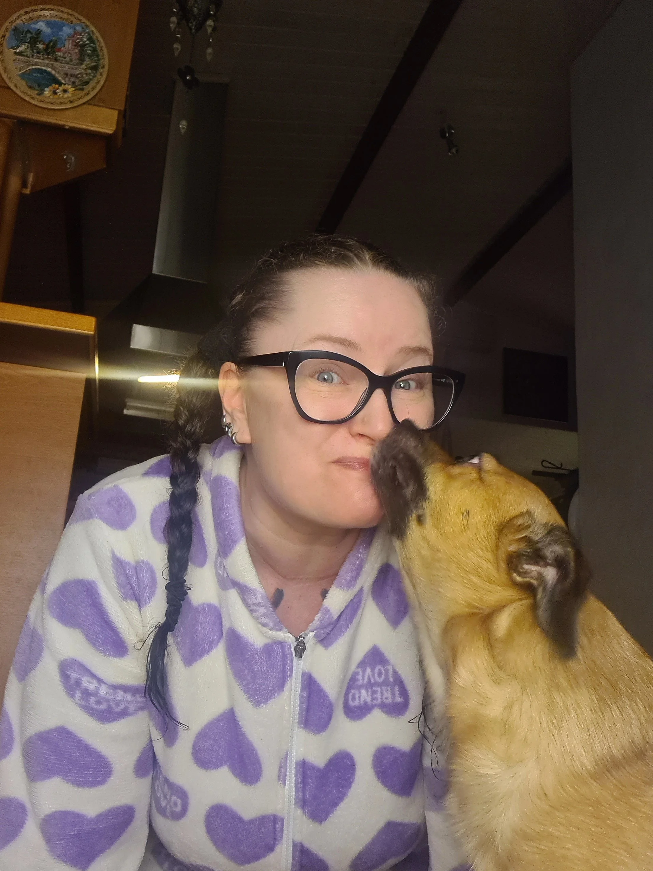 A woman wearing glasses and a white fleece jacket with purple hearts, holding a puppy against her face in a room with dark ceilings and a chandelier.