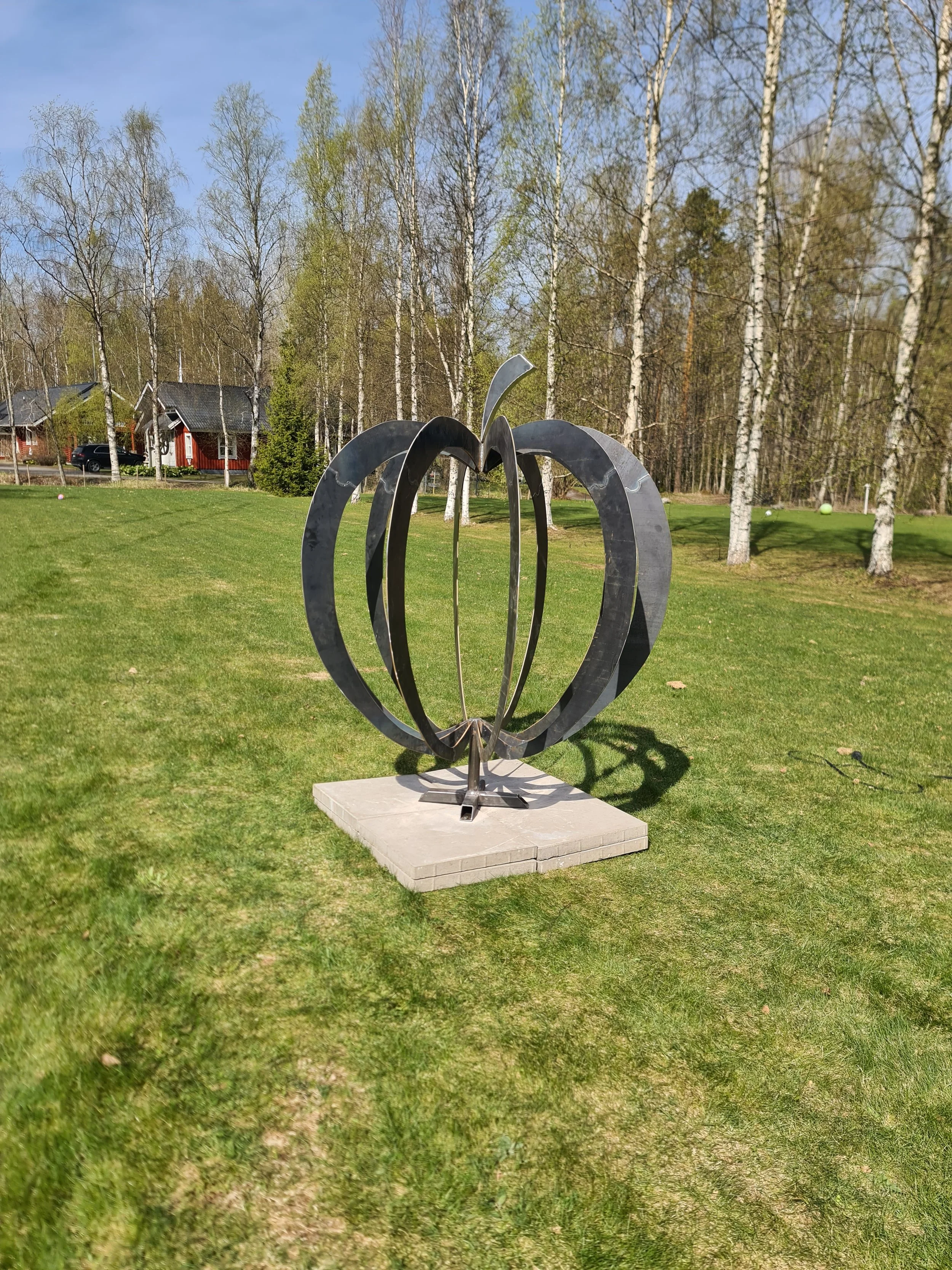 Modern metal sculpture resembling a tulip with open petals, mounted on a concrete platform, set in a grassy park area with trees and houses in the background.