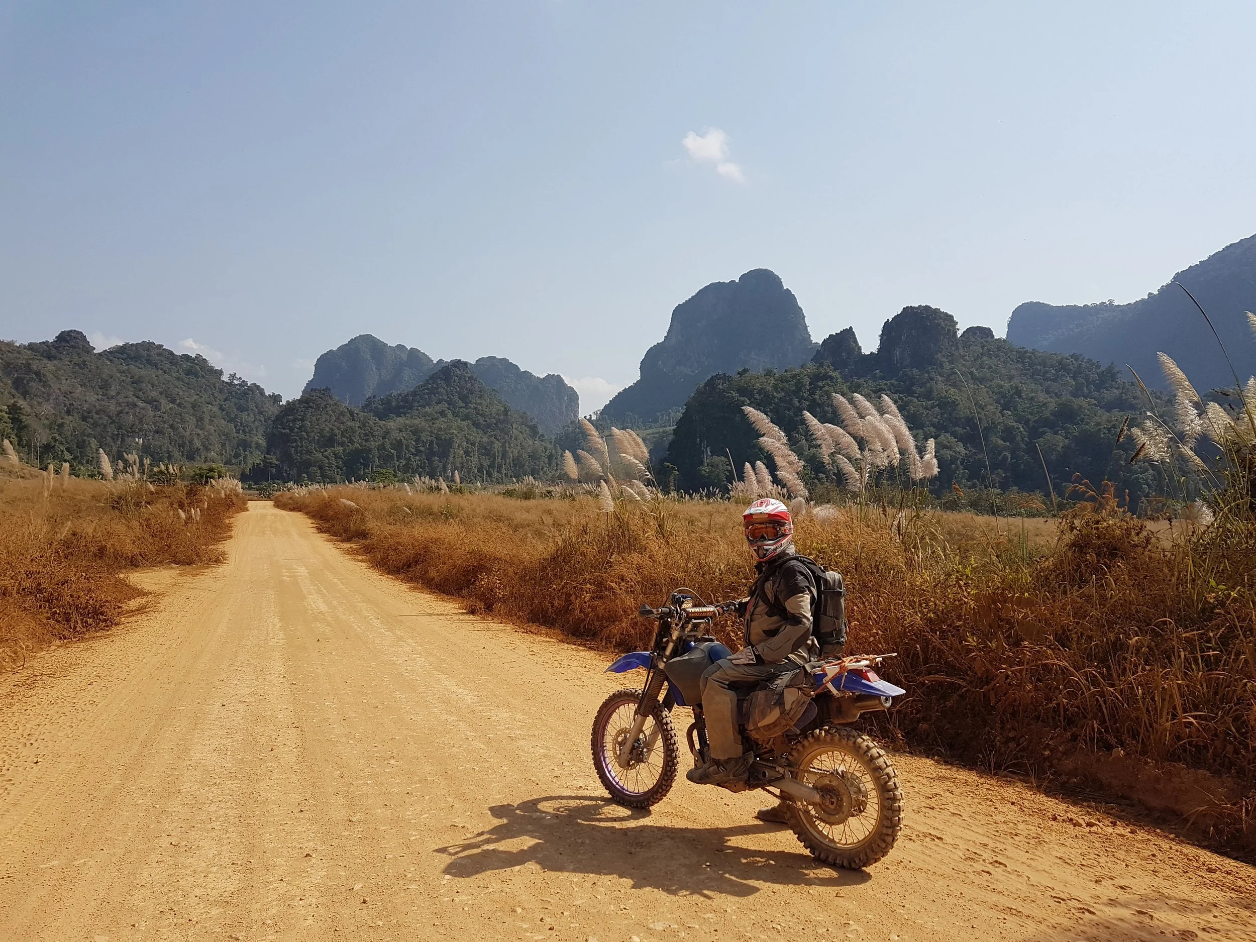Un motard en tenue adaptée, avec casque, assis sur une moto tout-terrain sur une route en terre dans un paysage de montagnes verdoyantes sous un ciel clair.