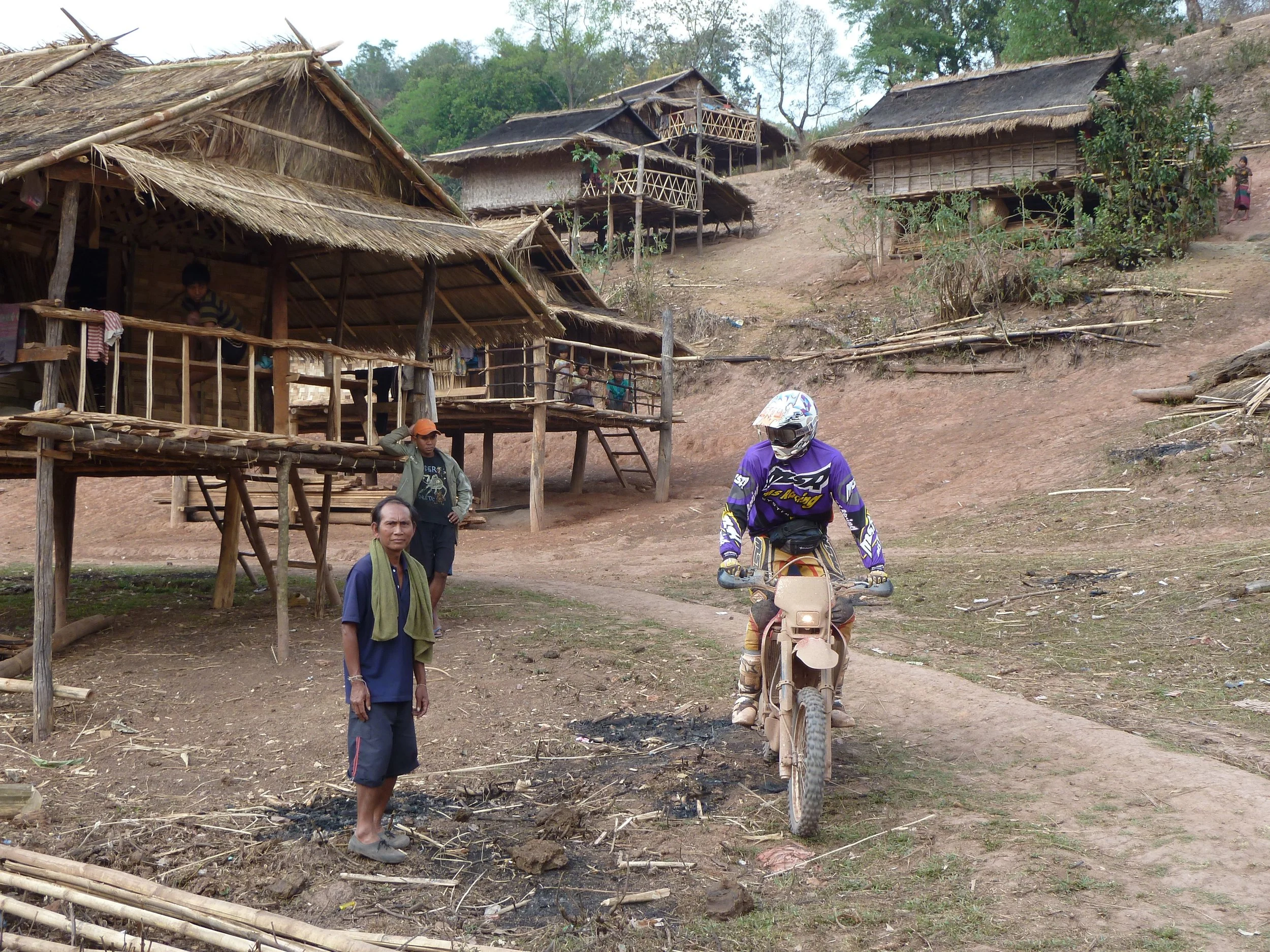 Hameaux en bois et bambou sur une pente, plusieurs personnes dont une sur une moto, dans un village rural avec végétation large et arbres.