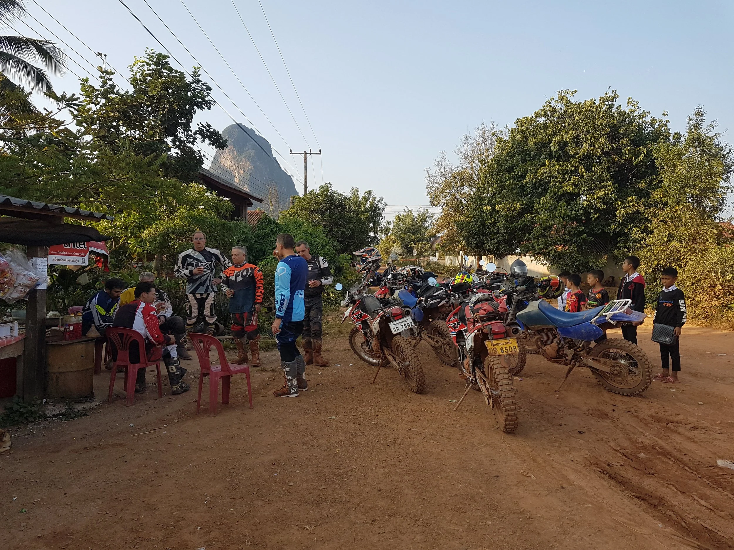Groupe de personnes et des motos dans une zone extérieure arborée, avec des montagnes en fond.