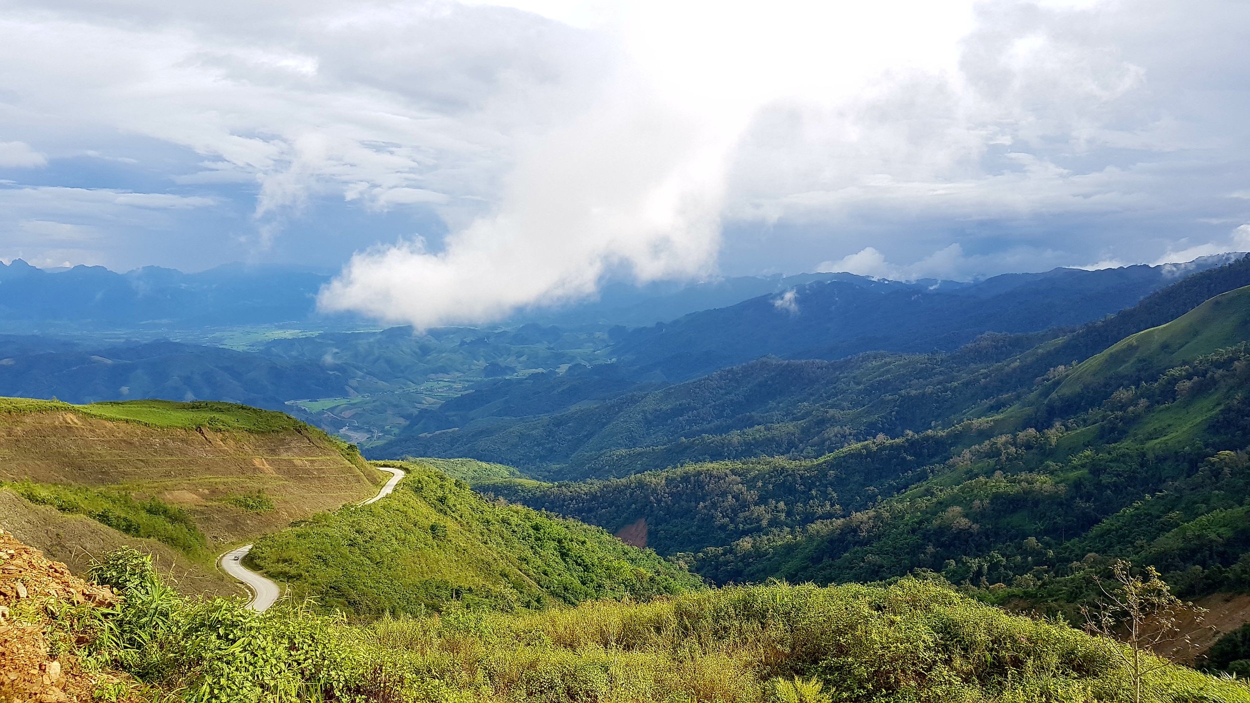 Paysage de montagnes verdoyantes avec un ciel nuageux, une route serpentant à travers la vallée