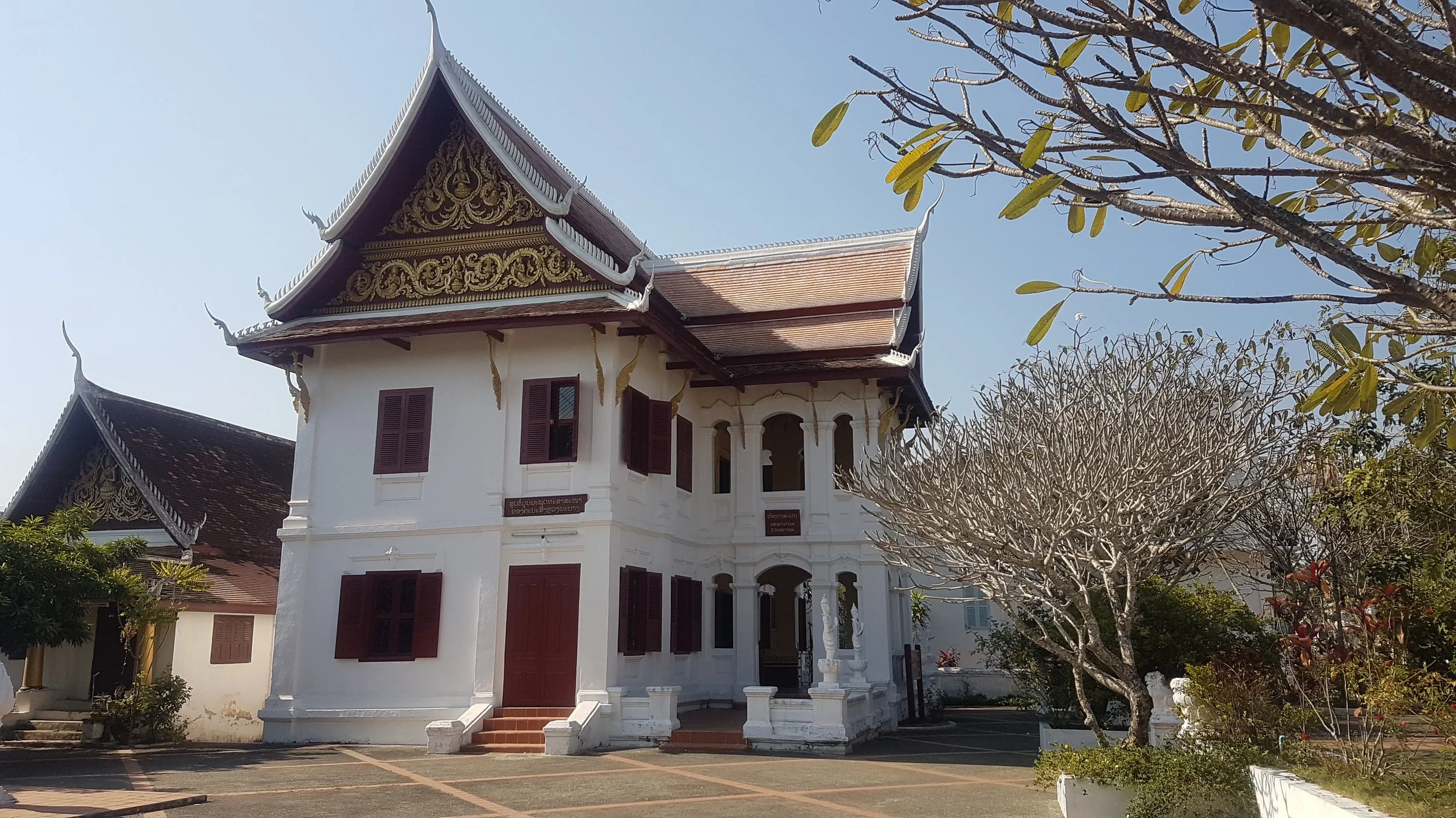 Une maison traditionnelle d'architecture asiatique avec un toit à plusieurs niveaux décoré de motifs dorés, entourée de végétation avec un arbre aux branches nues et un ciel bleu clair. Luang Prabang Laos.
