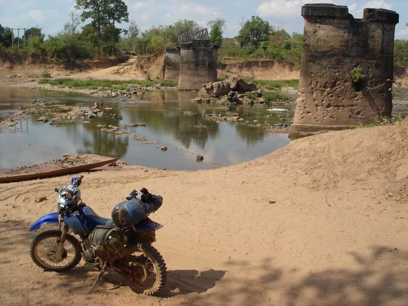 Une moto bleu avec des sacs attachés, garée sur un sol sableux près d'une rivière, avec des ruines ou un pont en pierre en arrière-plan. Le ciel est en partie nuageux. Au Laos.