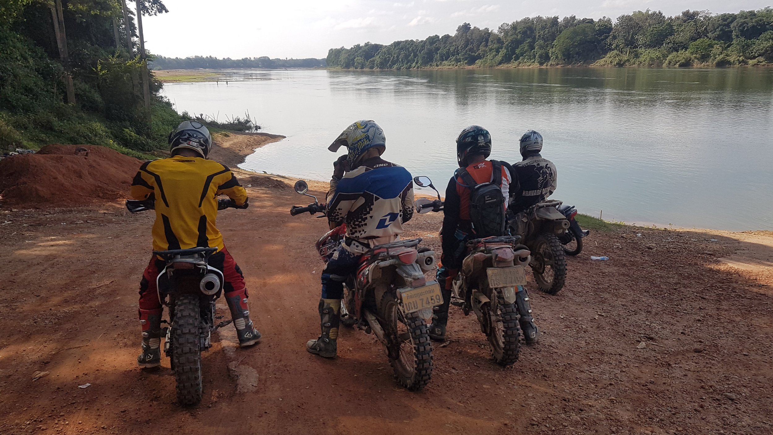 Quatre motocyclistes avec équipements de protection assis sur leurs motos, regardant une rivière côté nature avec des arbres verdoyants, par une journée ensoleillée.