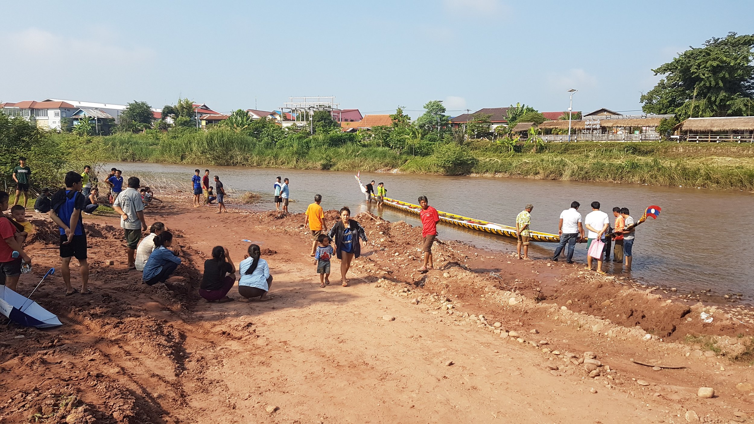 Groupe de personnes au bord d'une rivière, avec un bateau décoré, en train de faire une activité liée à la navigation ou à un événement culturel, dans un environnement rural.