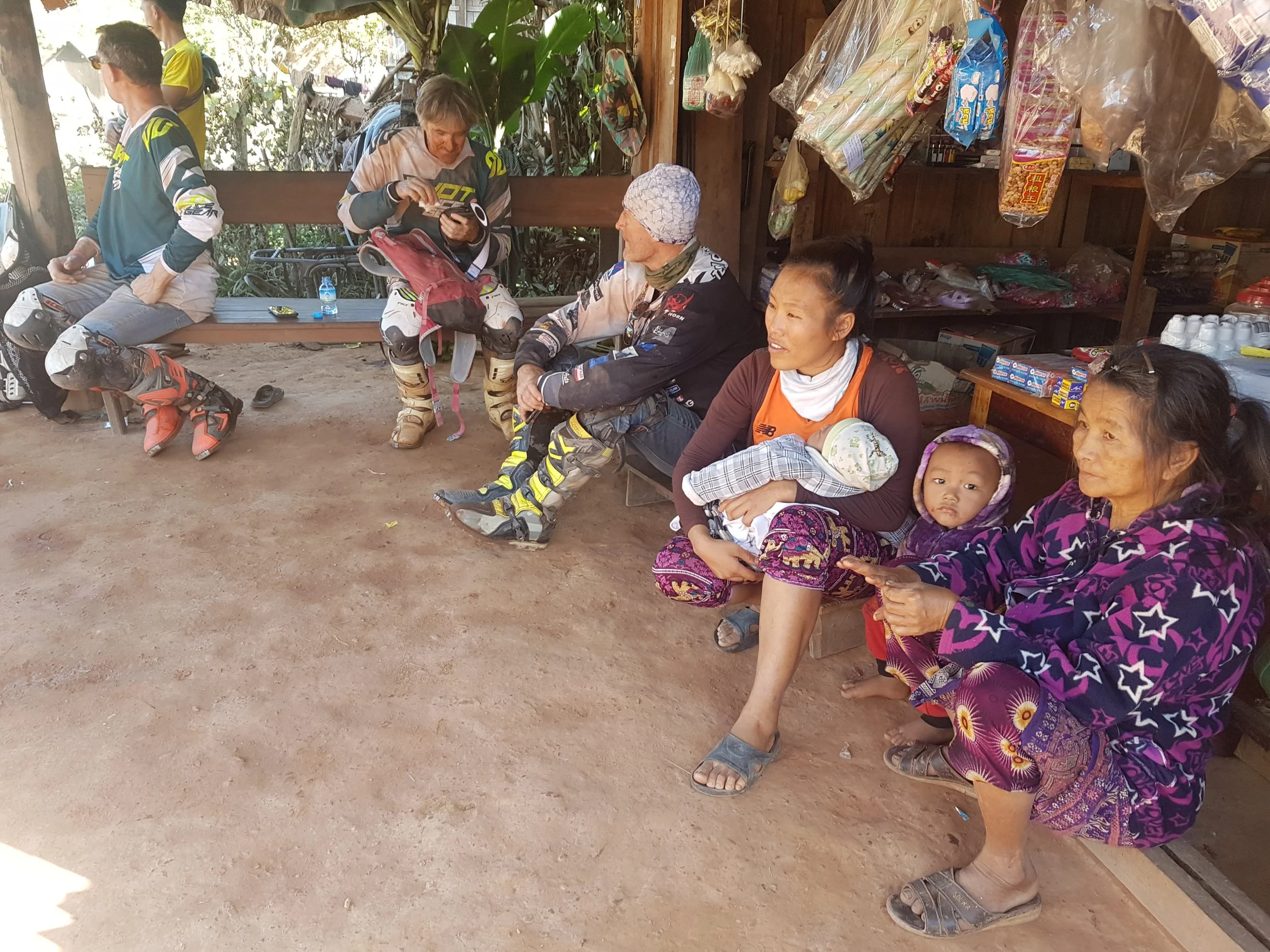 Groupe de personnes assises sur une banquette en bois à l'intérieur d'une petite boutique ou stand, avec des produits d'épicerie en plastique accrochés au mur derrière eux, des enfants, des femmes et un homme portant des vêtements de motocross, dans 