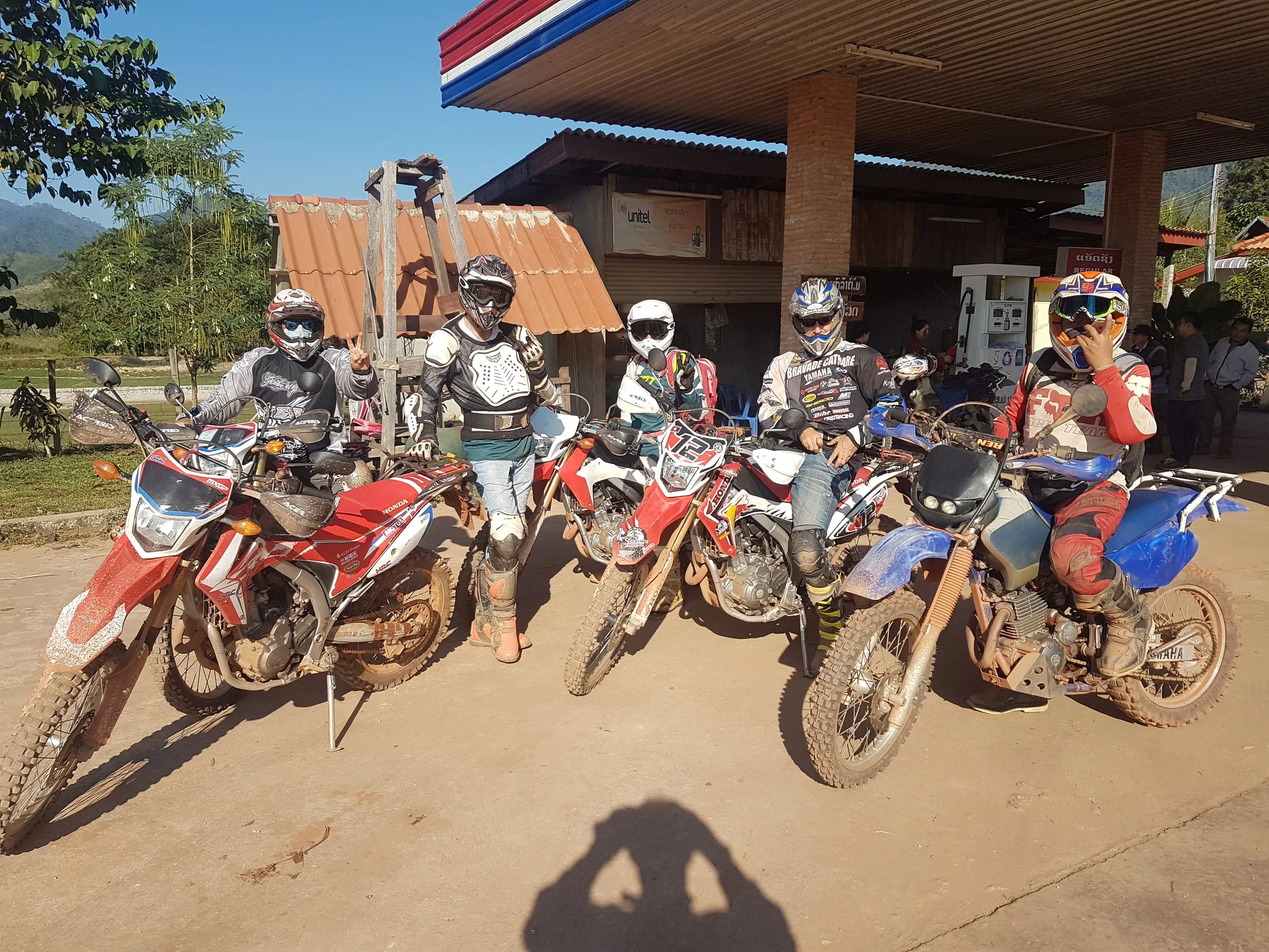 Groupe de motards portant des casques et des vêtements de protection, posant avec leurs motos tout-terrain, devant un bâtiment dans une région montagneuse.