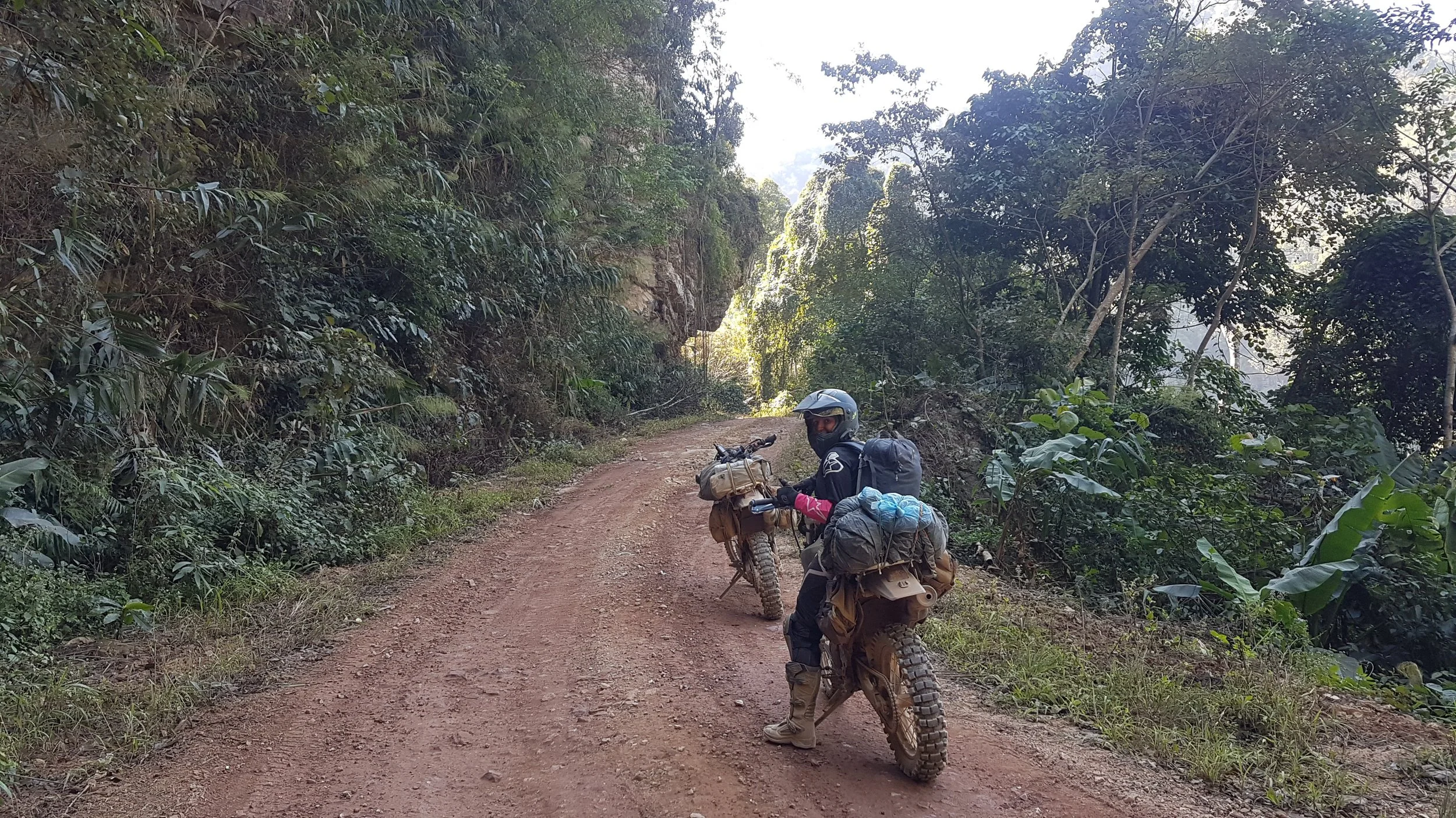 Une personne en tenue de moto tout-terrain, portant un casque, se trouve à côté d'une moto chargée, sur un chemin en terre dans une forêt luxuriante.