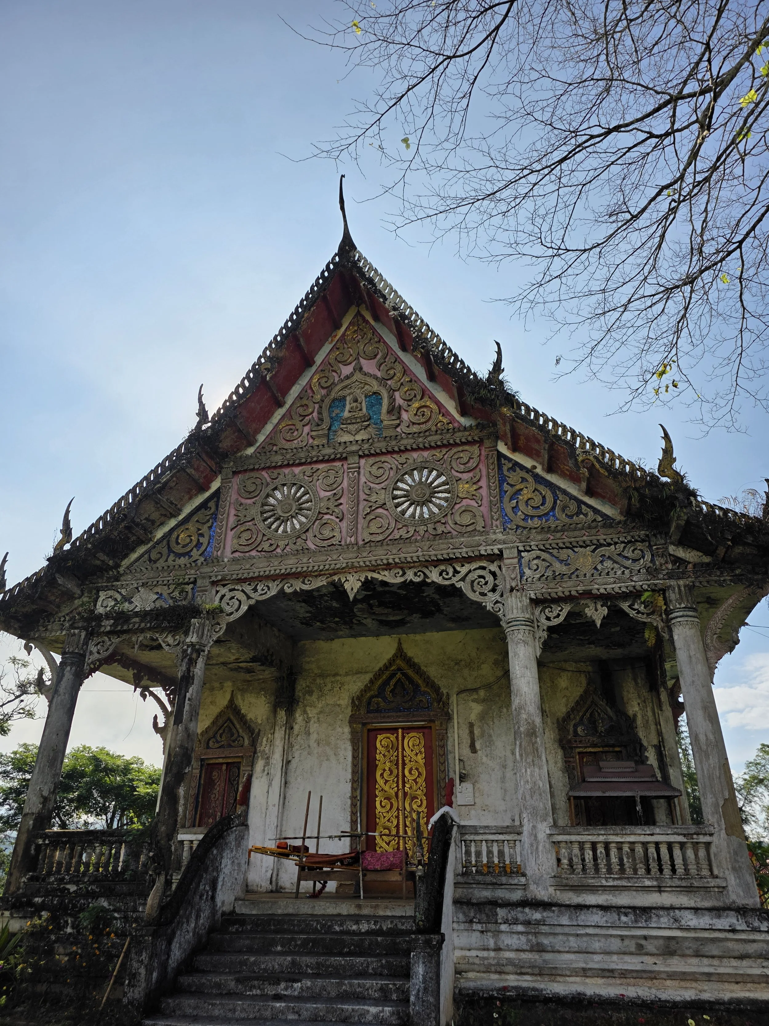 Une ancienne structure en bois et en pierre avec un toit à pointe, ornée de décorations traditionnelles, entourée d'arbres sans feuilles et un ciel bleu en arrière-plan.