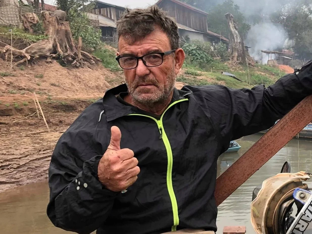 Homme portant un manteau noir avec un zip jaune, faisant un pouce en l'air, devant un paysage de rivière avec des arbres abattus et de la fumée en arrière-plan.