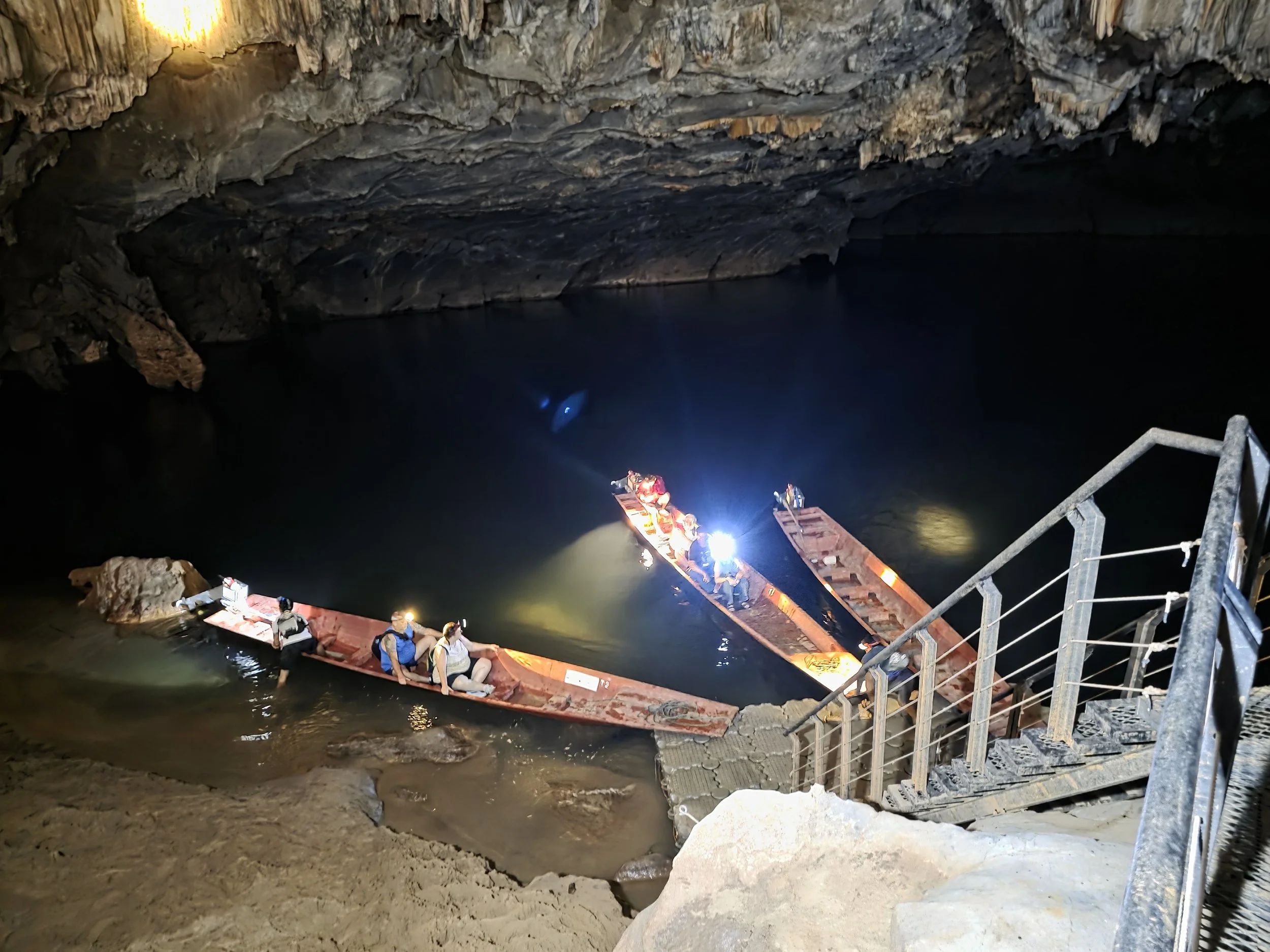 Une grotte avec un lac, un groupe de personnes en bateau éclairé par des lumières, vue depuis l'entrée de la grotte avec des escaliers en métal.