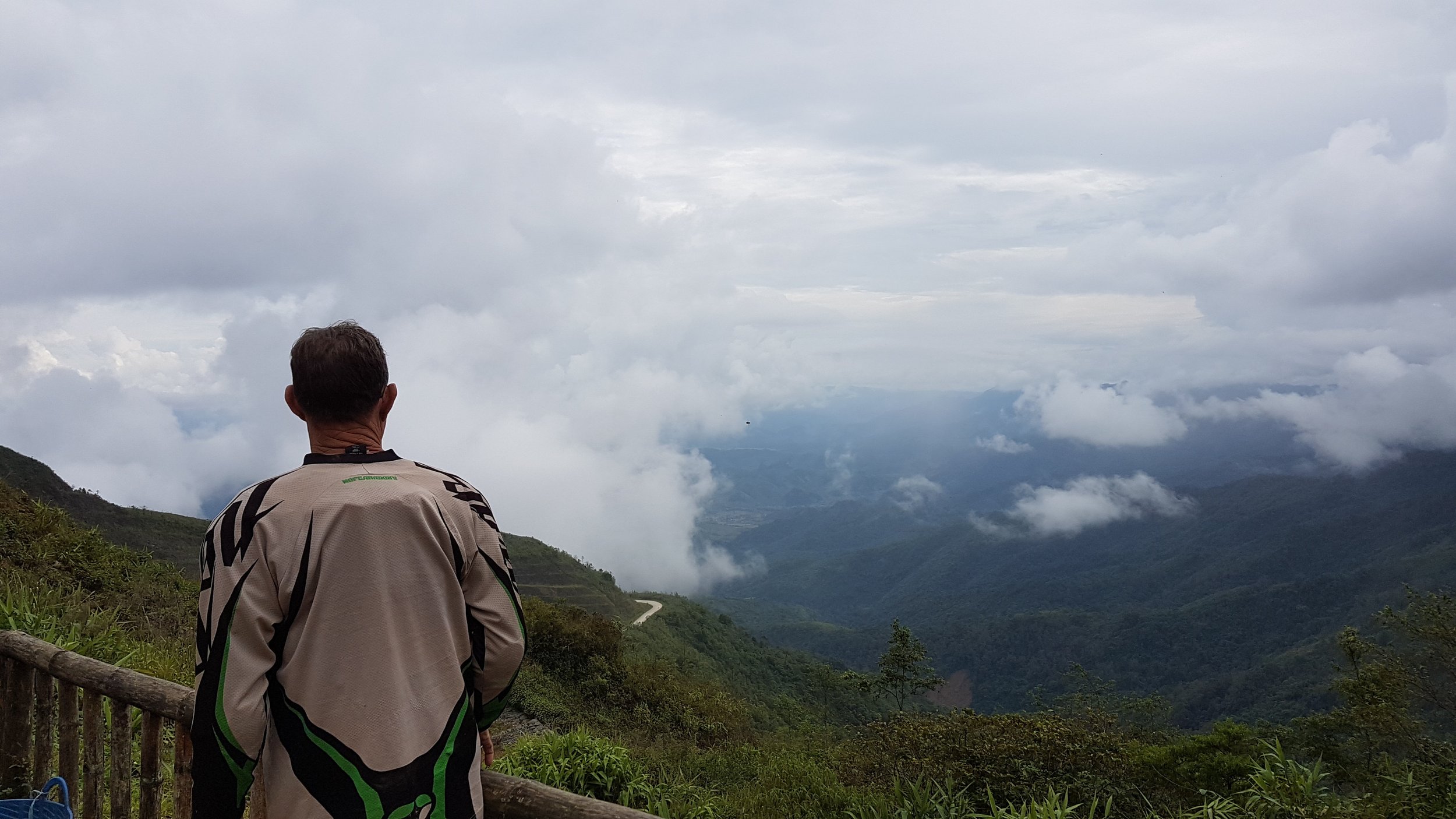 Un homme assis regardant un paysage de montagnes enveloppées de nuages, avec un sentier serpentant dans la vallée.