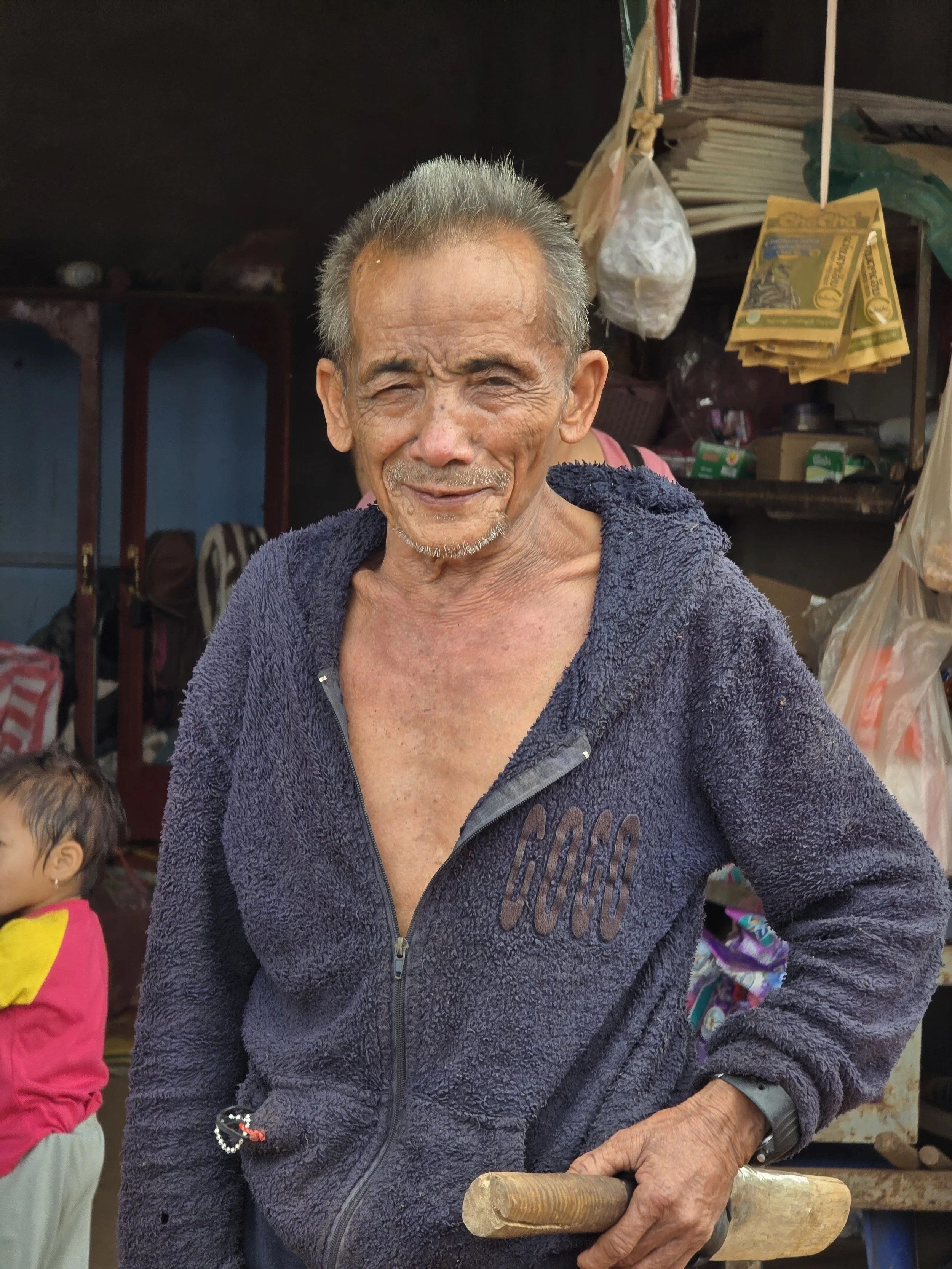 Un homme âgé souriant, portant une veste en polaire foncée, tient un outil en bois dans la main, dans un environnement intérieur avec des marchandises suspendues et des étagères.
