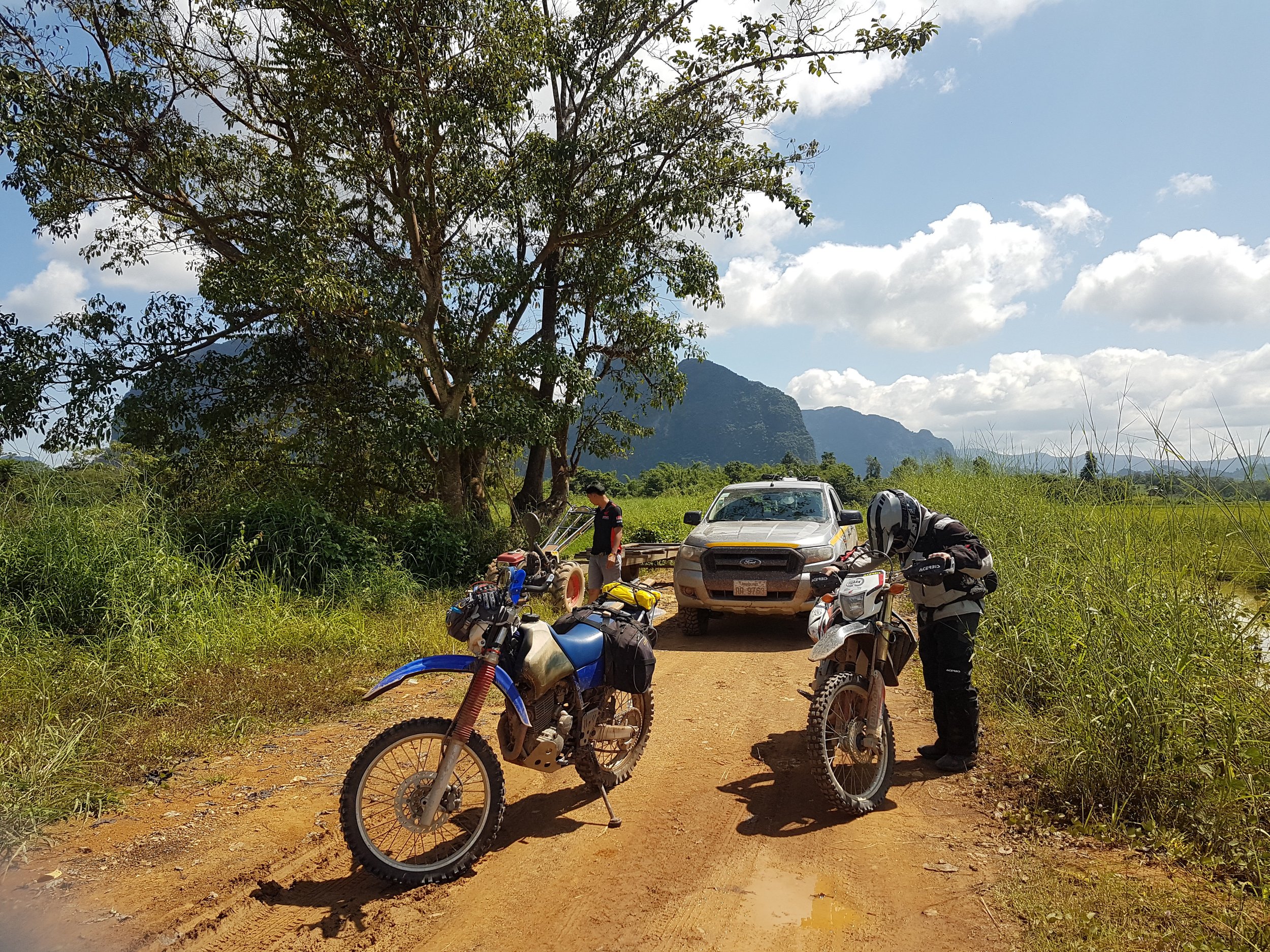 Le paysage montre une zone rurale avec des motos, une voiture, des personnes préparant des équipements, une grande étendue d'herbe, un arbre, des montagnes en arrière-plan et un ciel partiellement nuageux.