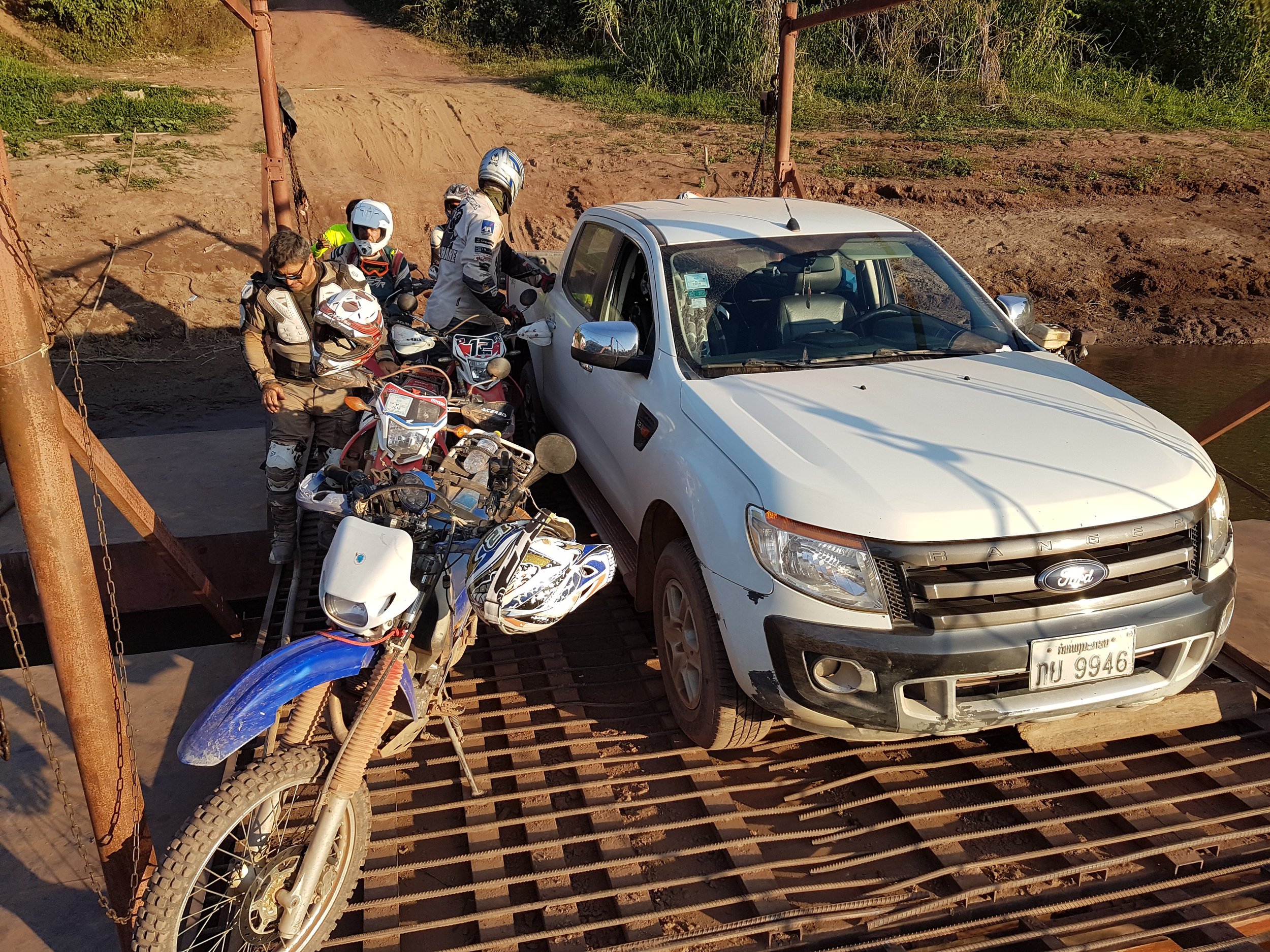 Un groupe de motards en tenue de protection attendaient avec des motos, une voiture blanche est arrêtée sur un pont en fer. L'environnement semble rural avec un chemin de terre en arrière-plan.
