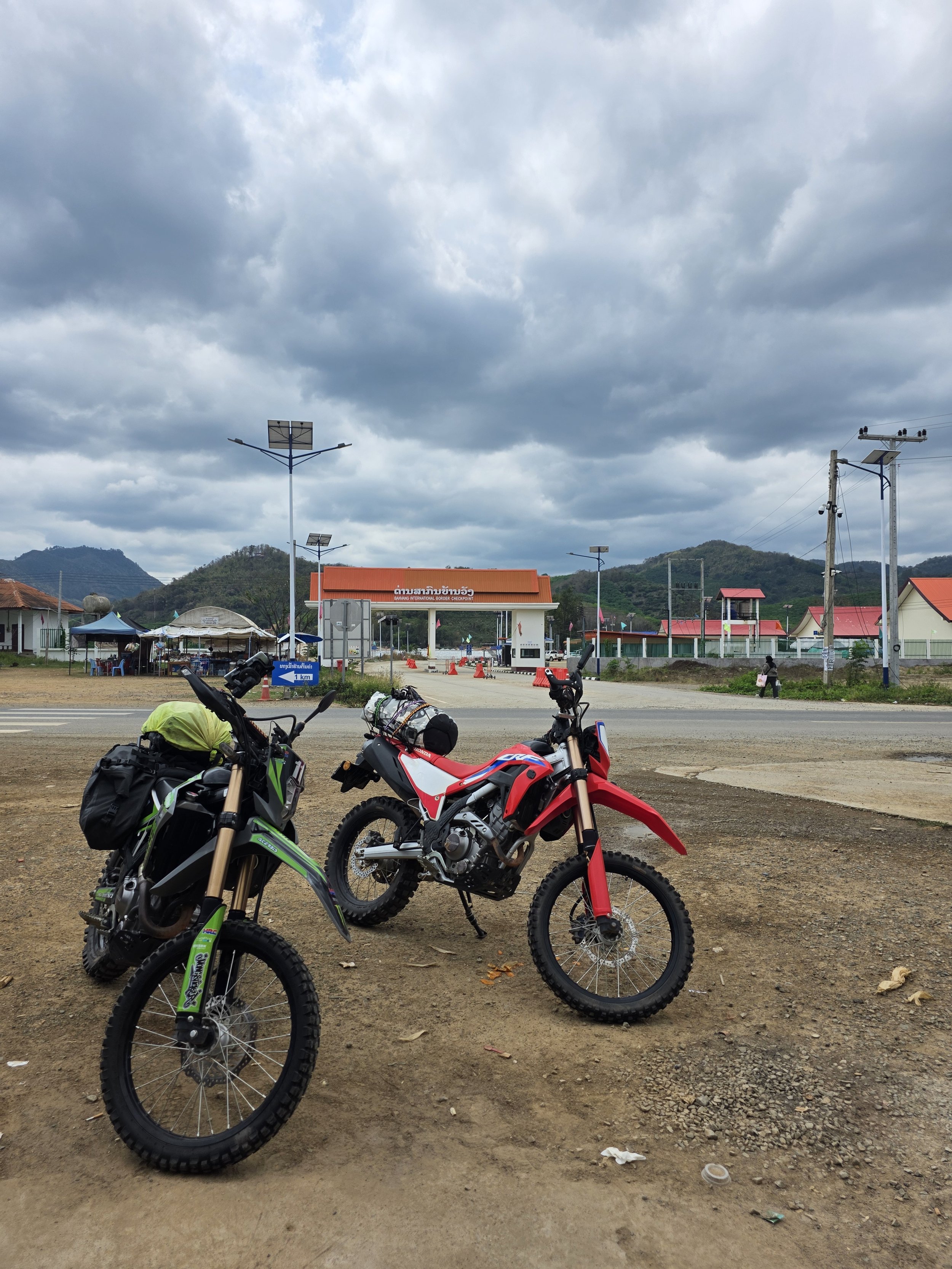 Deux motos tout-terrain (motocross) vert et rouge garées sur un terrain en terre, avec une frontière à l'entrée d'un poste frontière ou contrôle, sous un ciel nuageux, avec des montagnes en arrière-plan.
