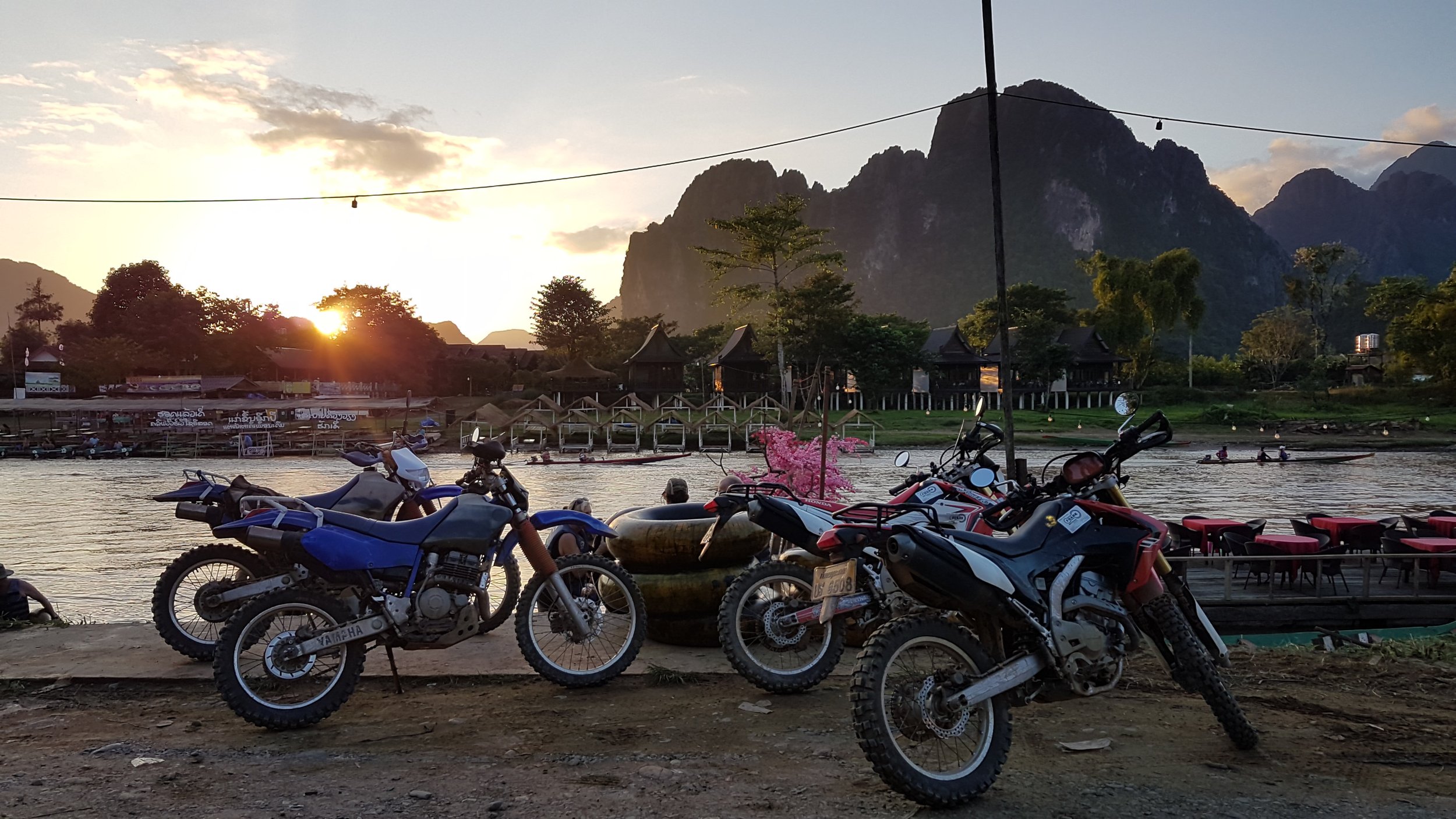Motocyclettes garées près d'une rivière au coucher du soleil, avec des montagnes en arrière-plan et des bateaux naviguant sur l'eau.