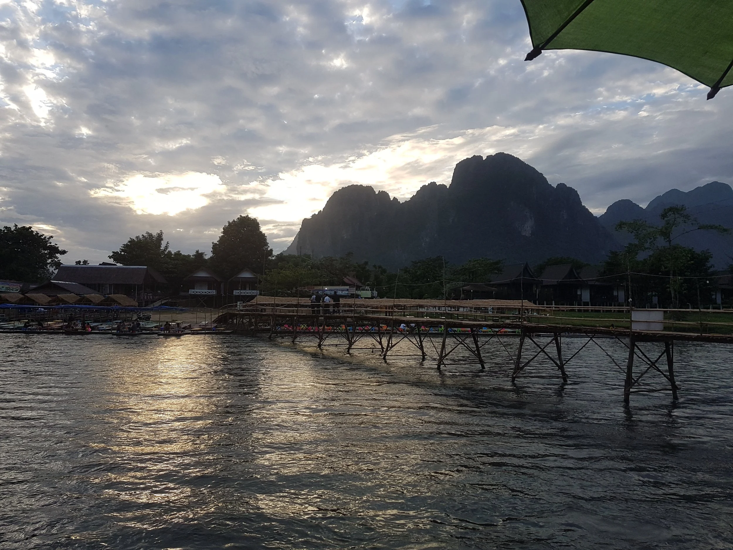 Un paysage de montagne avec un ciel nuageux et un coucher de soleil, vu depuis une zone boisée avec des maisons et une rivière en premier plan.