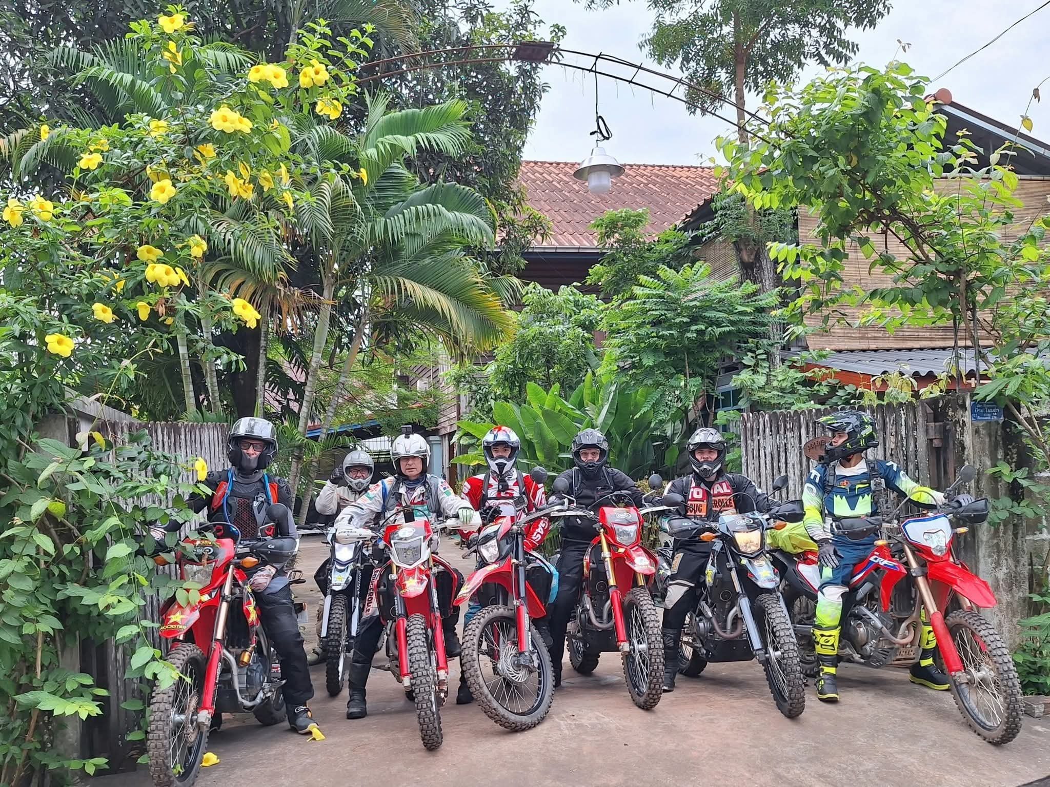 Groupe de motards avec des casques et des motos rouges, posant devant une maison avec un toit en tuiles, entourés de végétation luxuriante et d'arbres.