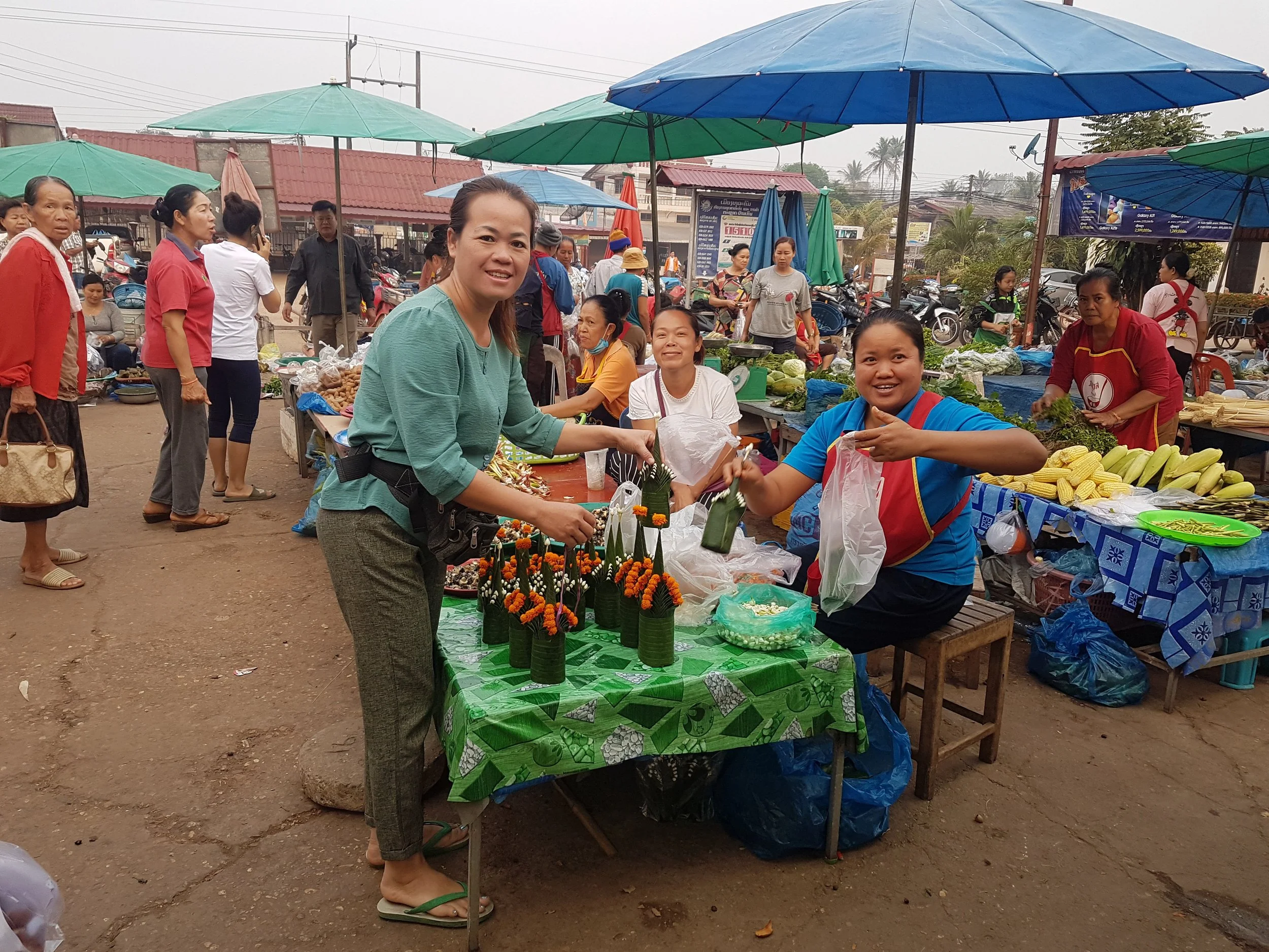 Un marché en plein air avec plusieurs stands vendant des légumes, des fleurs, et d'autres produits, sous des parasols colorés. Des personnes achètent et vendent dans une ambiance animée.