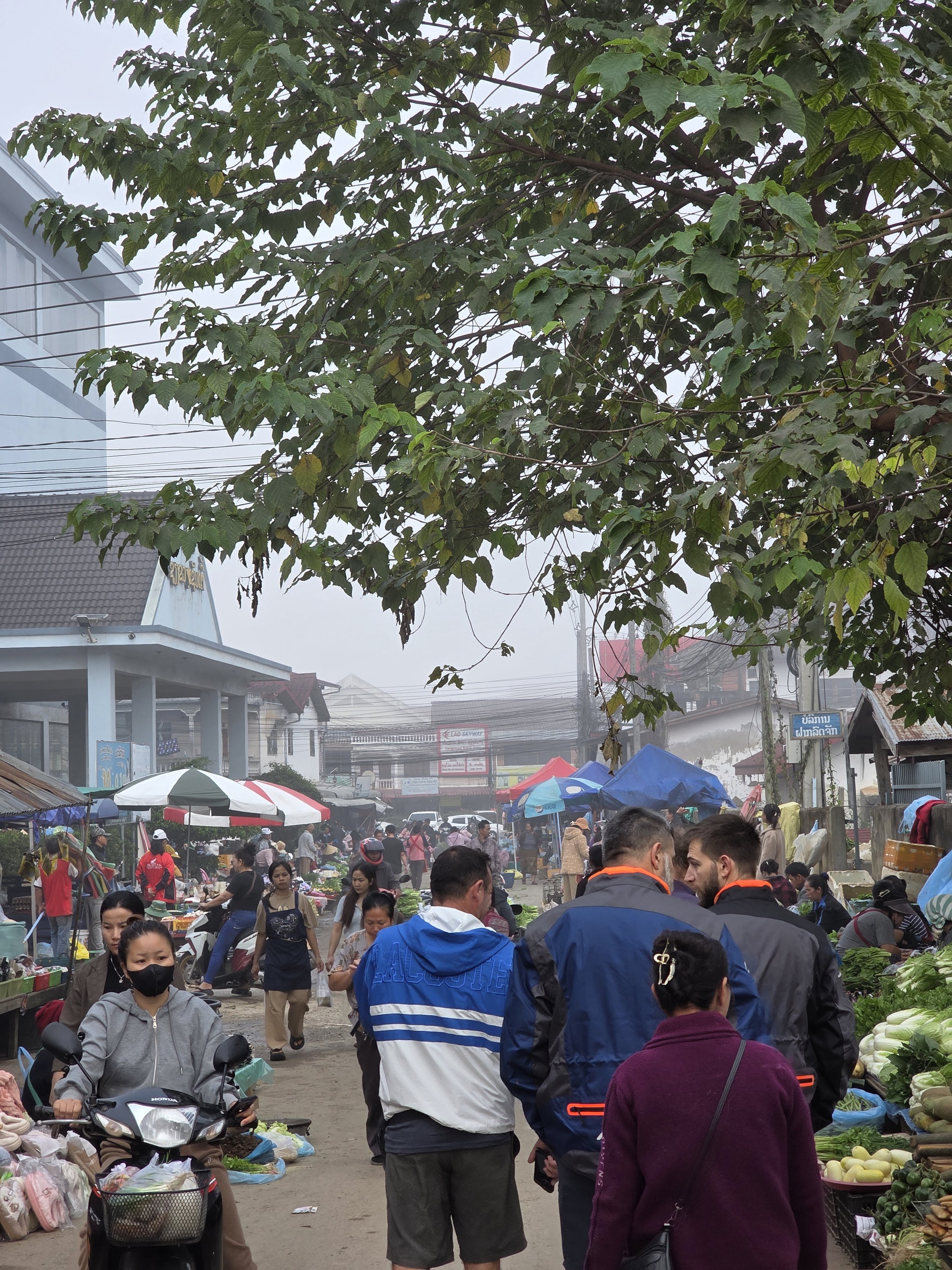 Un marché en plein air avec de nombreuses personnes achetant des légumes et d'autres produits, sous des parasols colorés, dans une ambiance animée et un ciel brumeux.