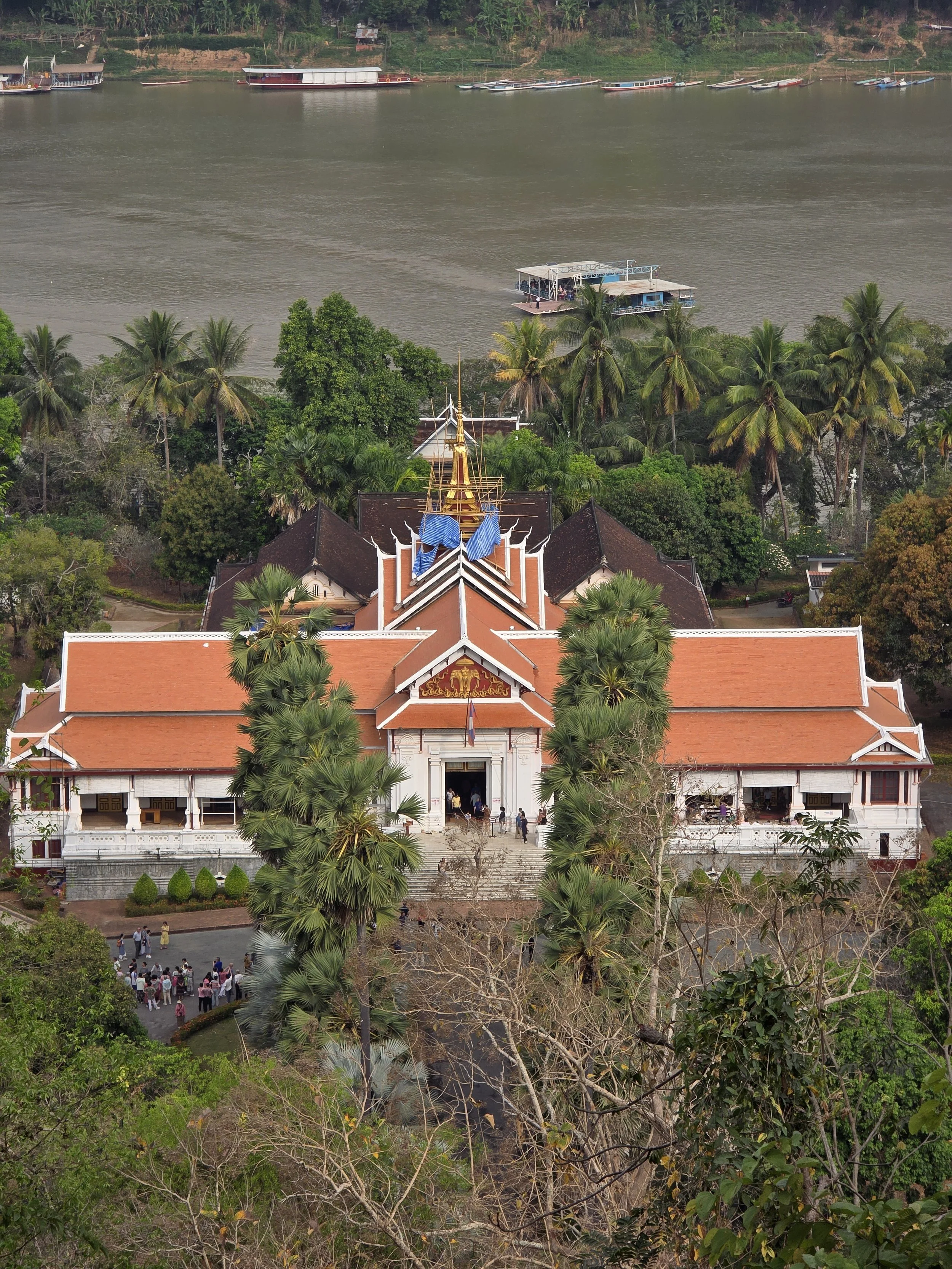 Vue aérienne d'un temple avec un toit rouge, entouré de palmiers, situé près d'une rivière où naviguent des bateaux. Des groupes de personnes se trouvent à l'entrée du temple et sur la place en face.