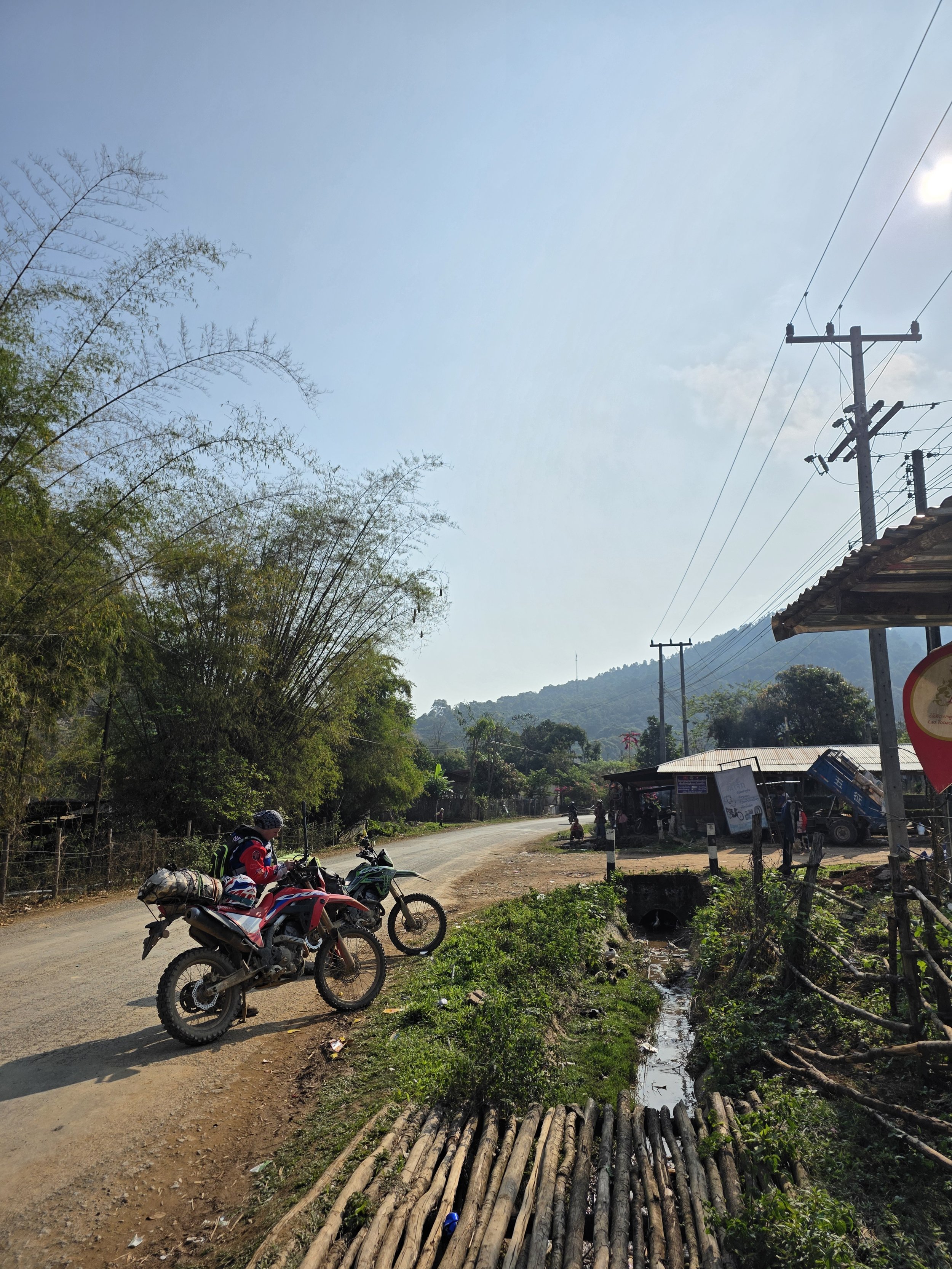 Une route rurale avec deux motards stationnés à côté, une petite boutique ou stand avec des personnes, plusieurs poteaux électriques avec lignes électriques, des arbres et une colline en arrière-plan, un ciel clair avec peu de nuages.
