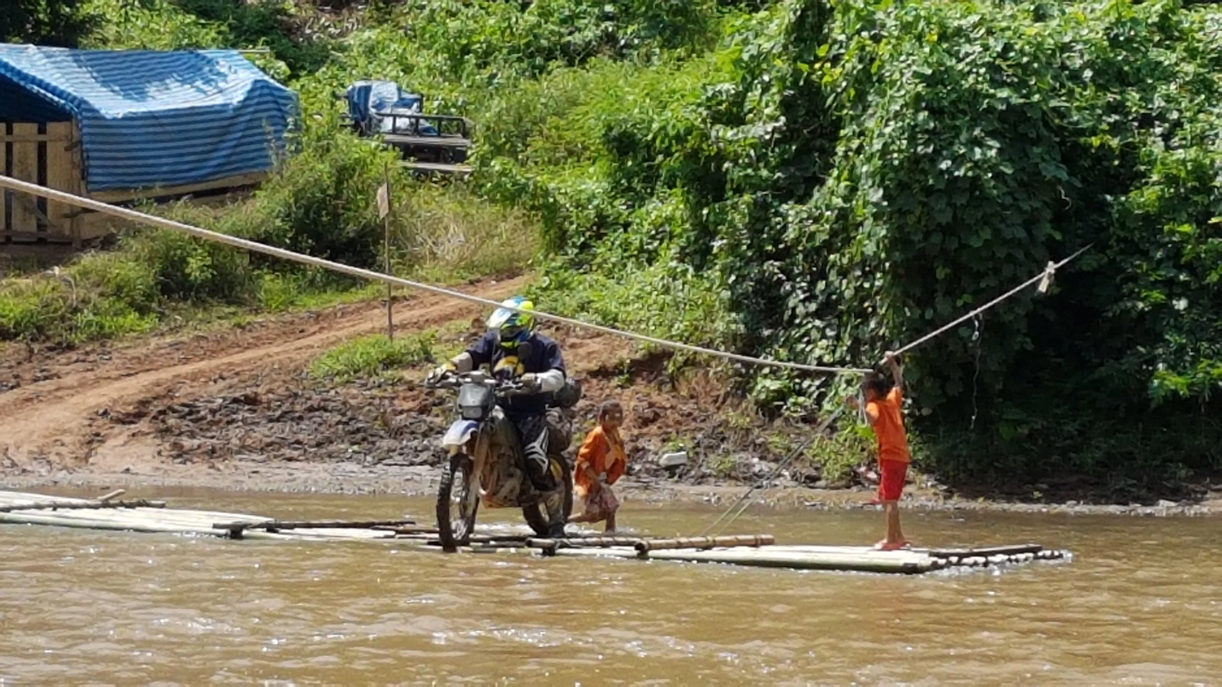 Personne en moto et deux enfants sur une berge près de l'eau, avec des arbres denses en arrière-plan.