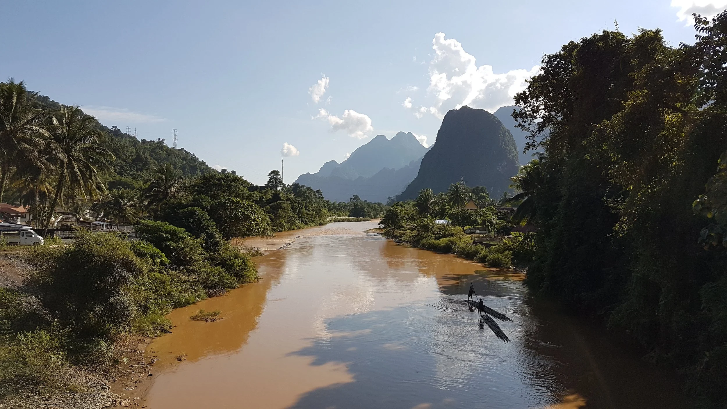 Une rivière marron traverse une région montagneuse et verte avec des arbres tropicals, des maisons et un bateau avec un pêcheur, sous un ciel en partie nuageux.