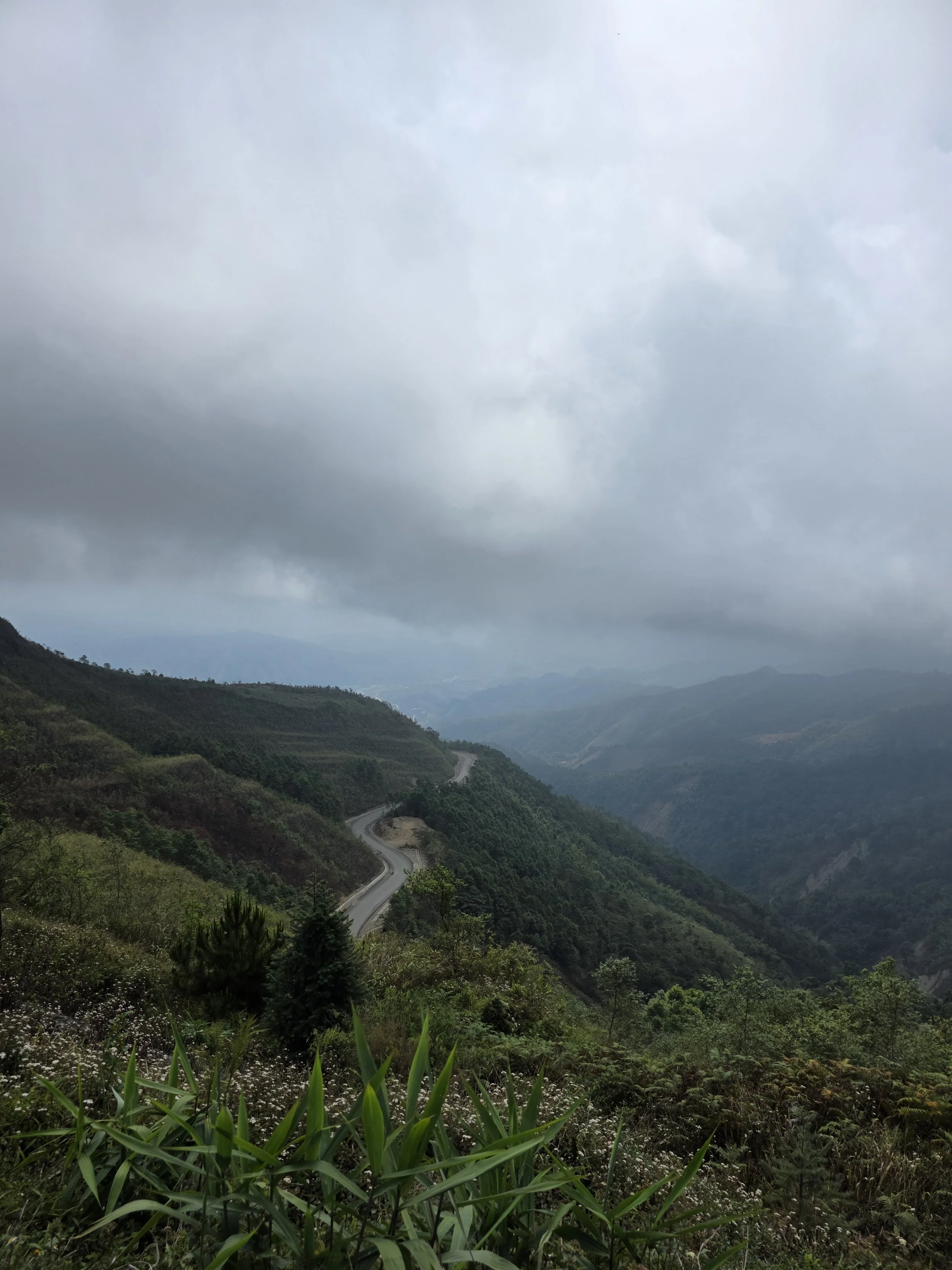 Paysage montagneux avec route serpentant à travers la végétation verdoyante sous un ciel nuageux.