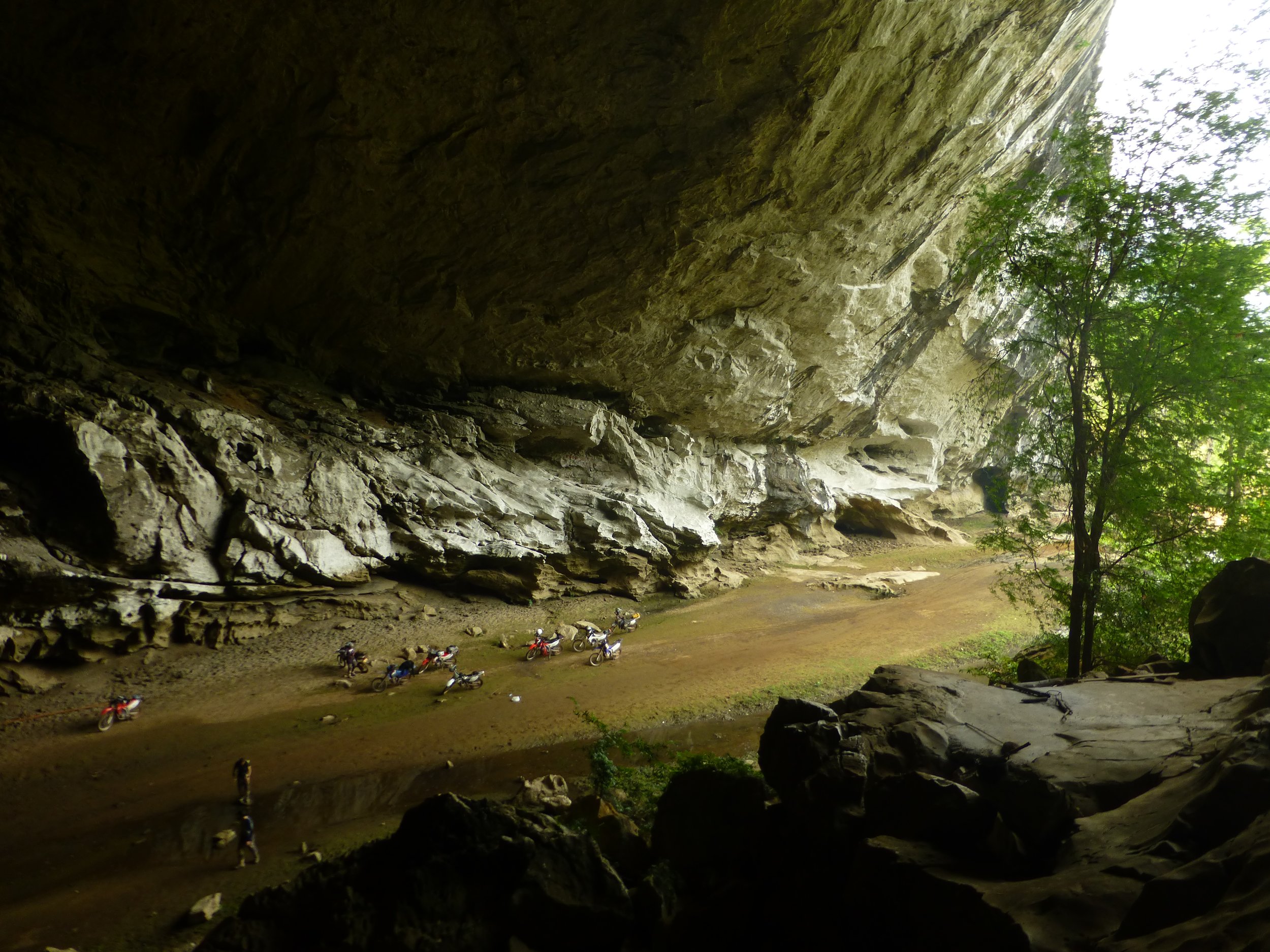 Une grotte avec une sortie en plein air laissant entrer la lumière, des arbres verts et plusieurs motos colorées stationnées à l'entrée.