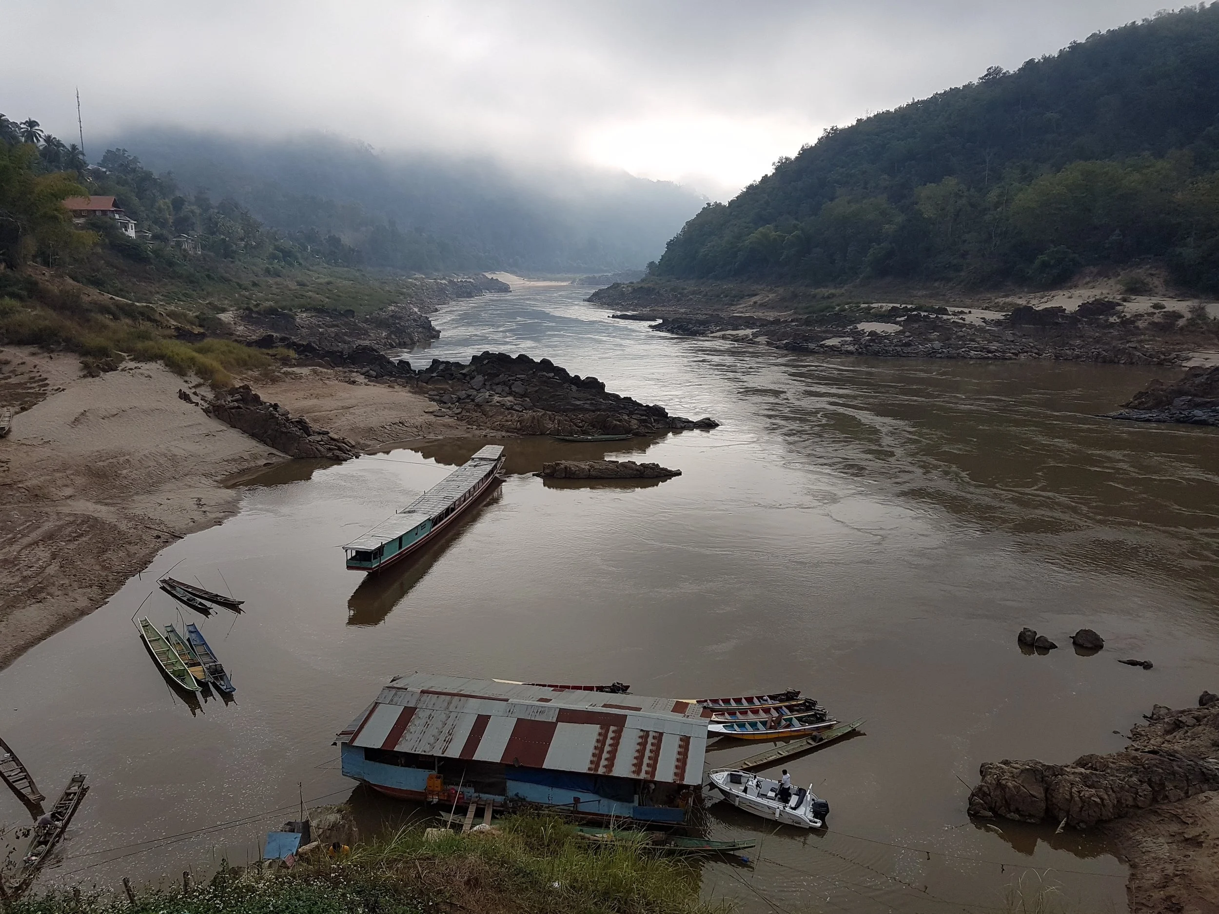 Vue d'une rivière entourée de montagnes verdoyantes sous un ciel brumeux, avec plusieurs bateaux amarrés près de la rive.