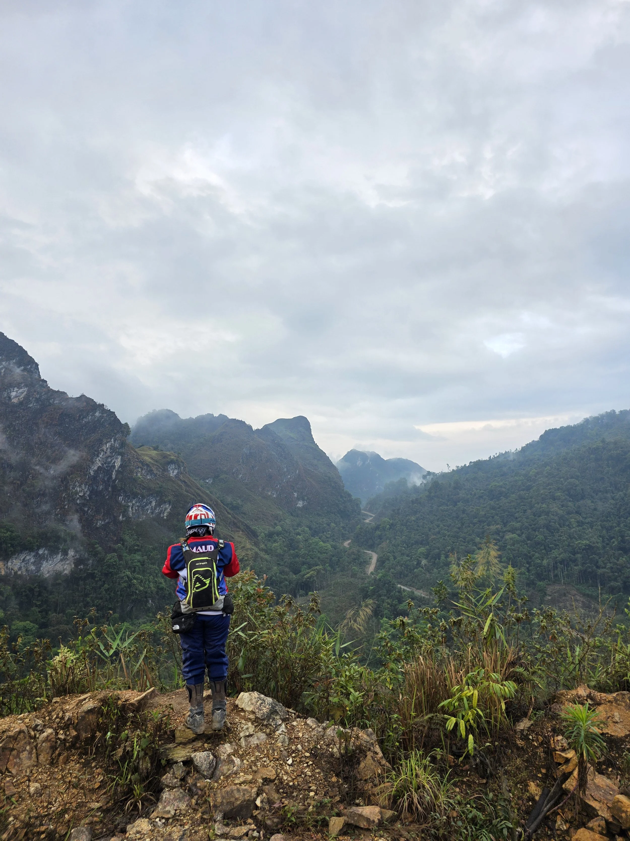 Une personne équipée de vêtements de randonnée et d'un casque, vue de dos, regarde un paysage de montagnes verdoyantes sous un ciel nuageux.