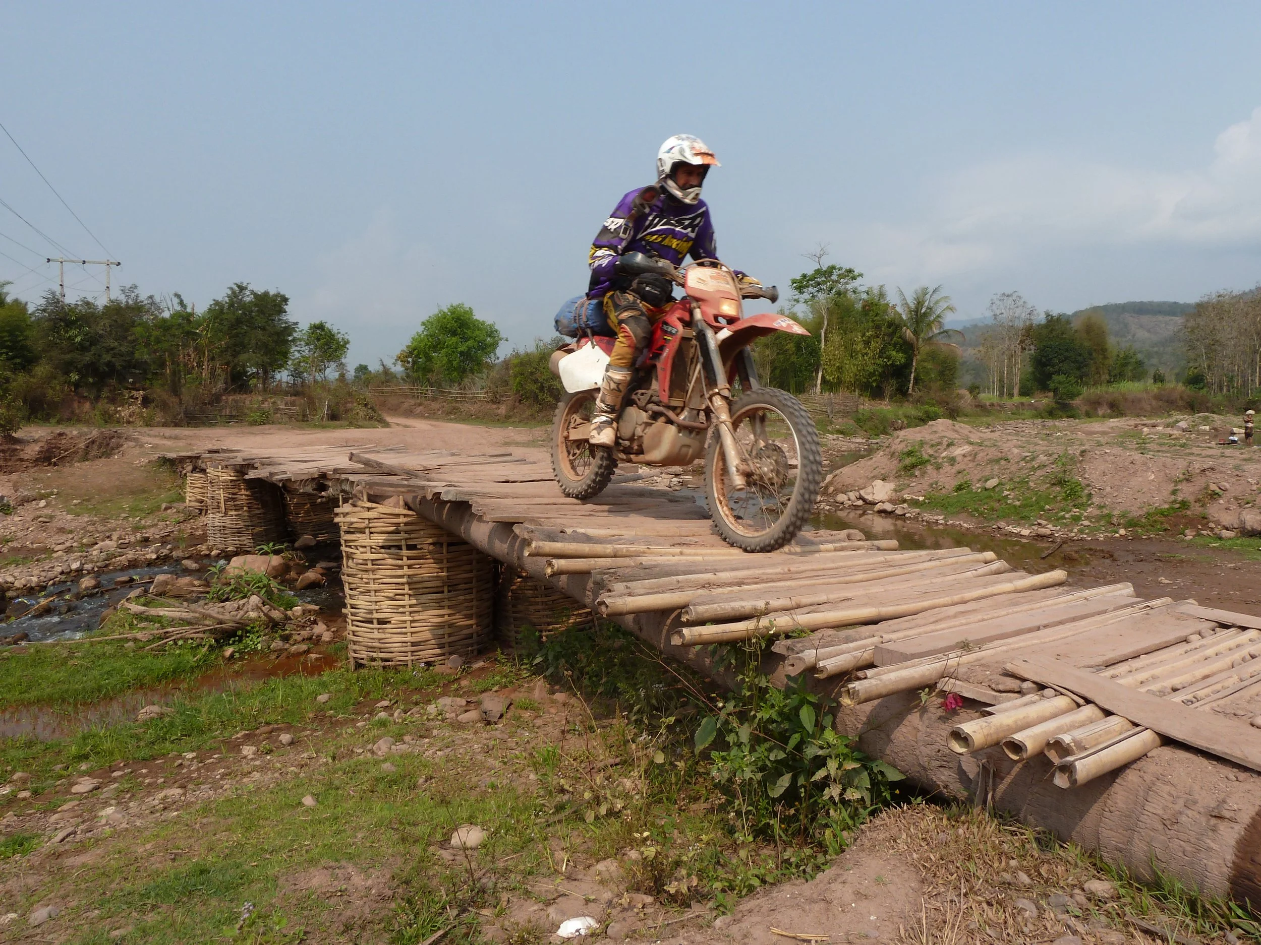 Un homme portant un casque et une tenue de motocross, chevauchant une moto rouge sur un pont en bois en milieu rural avec des arbres et un ciel partiellement nuageux.