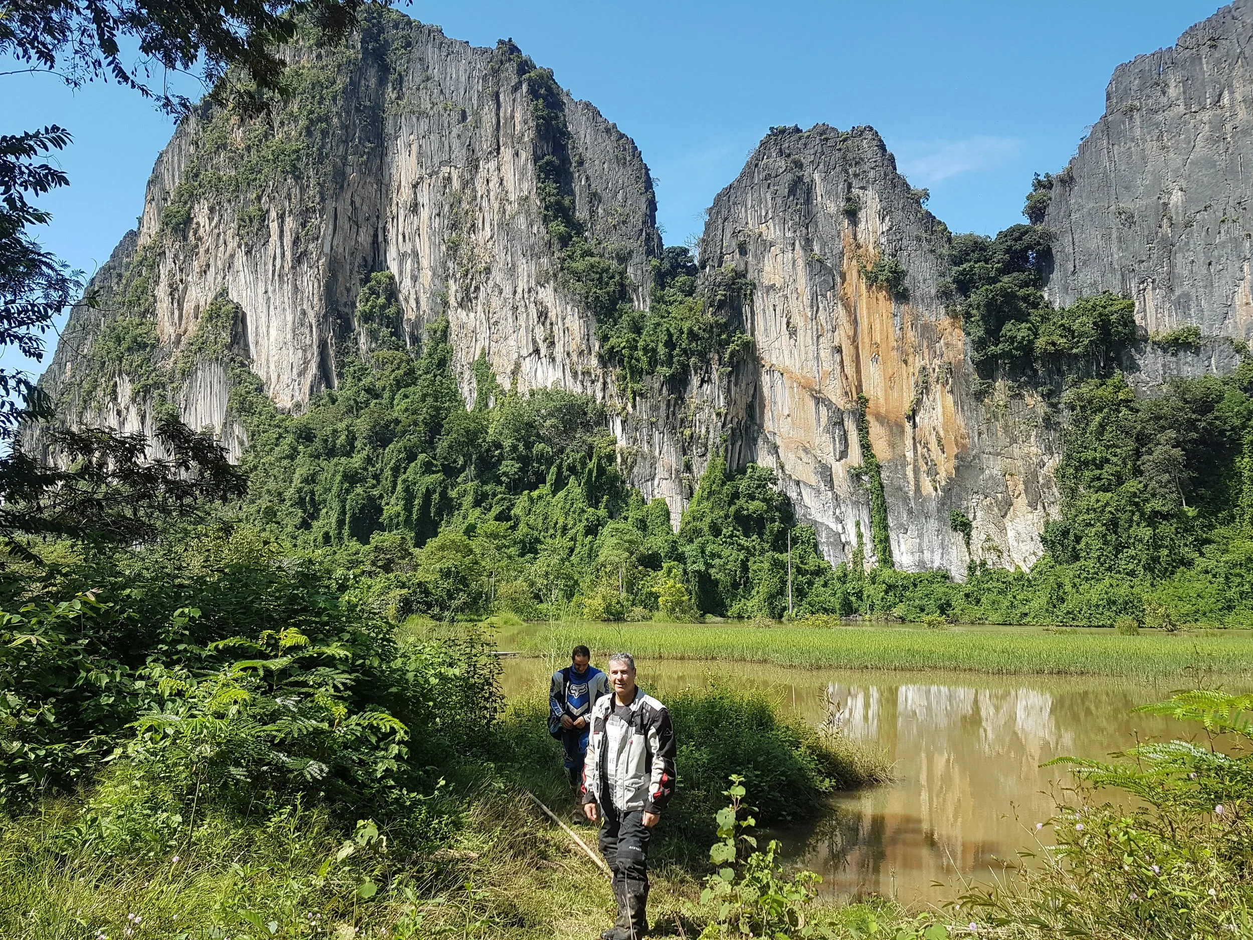 Deux personnes marchant le long d'une rivière entourée de végétation dense, avec d'imposantes formations rocheuses ou montagnes en arrière-plan sous un ciel bleu clair.