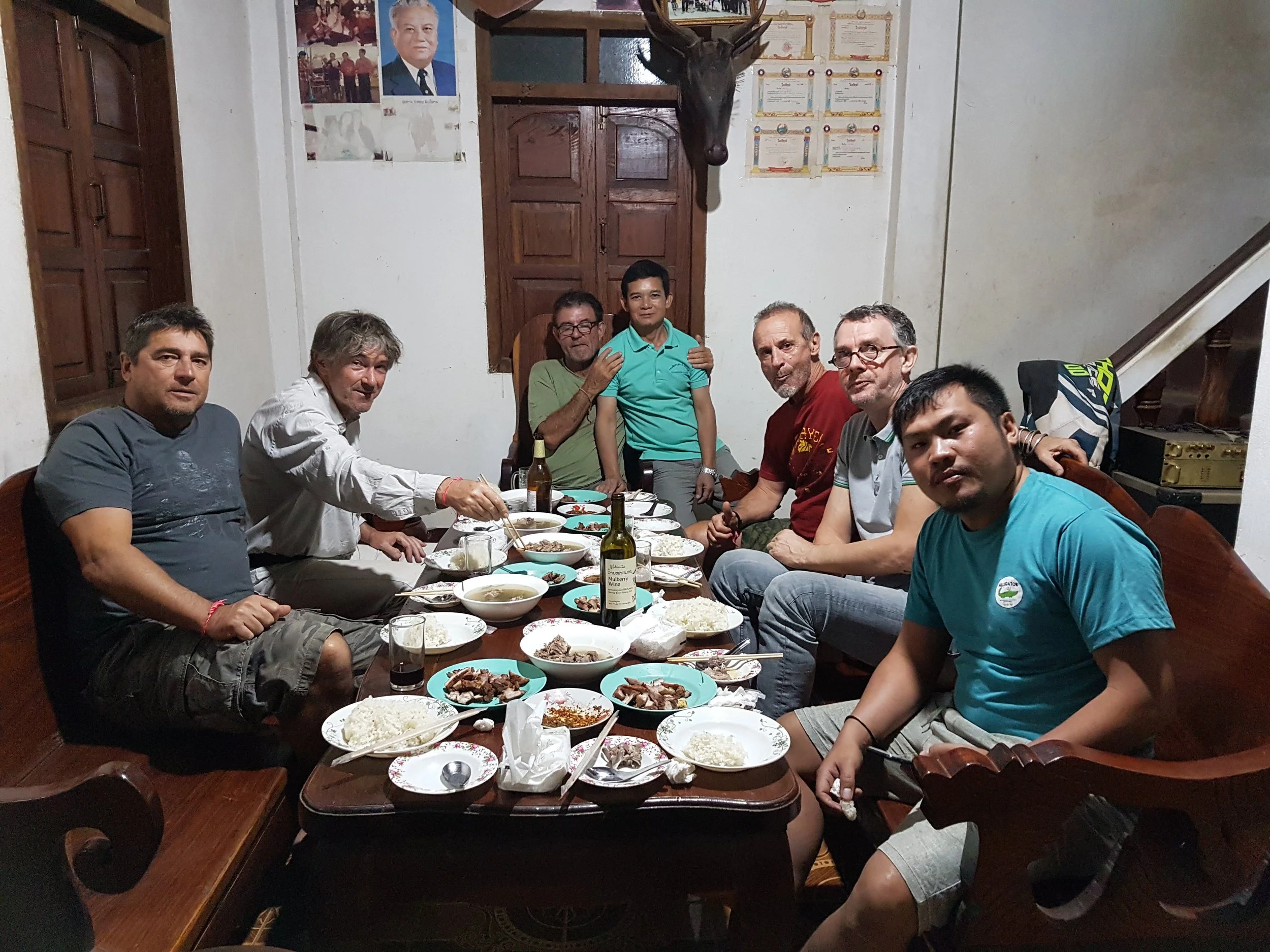 Groupe d'hommes assis autour d'une table avec de la nourriture, partageant un repas dans une pièce avec des diplômes et un portrait accroché au mur, une tête de cerf suspendue au mur