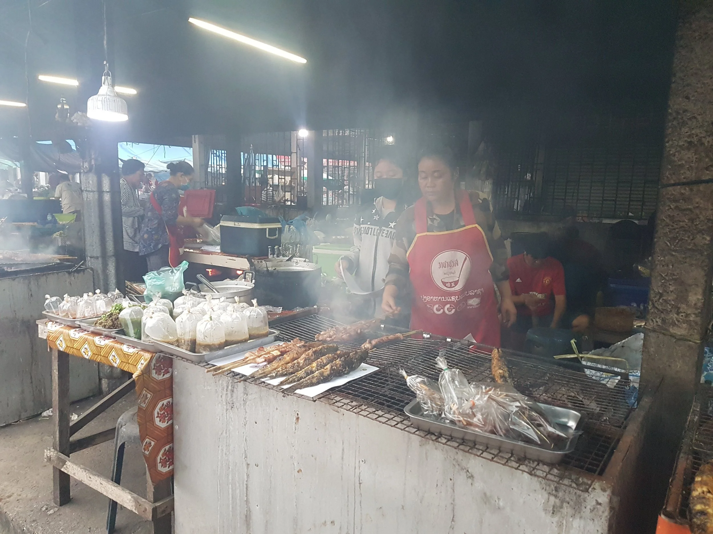 Un étal de barbecue avec des morceaux de viande grillés et des brochettes, dans un marché de nuit, avec des vendeuses en arrière-plan et des sacs en plastique contenant d'autres aliments.
