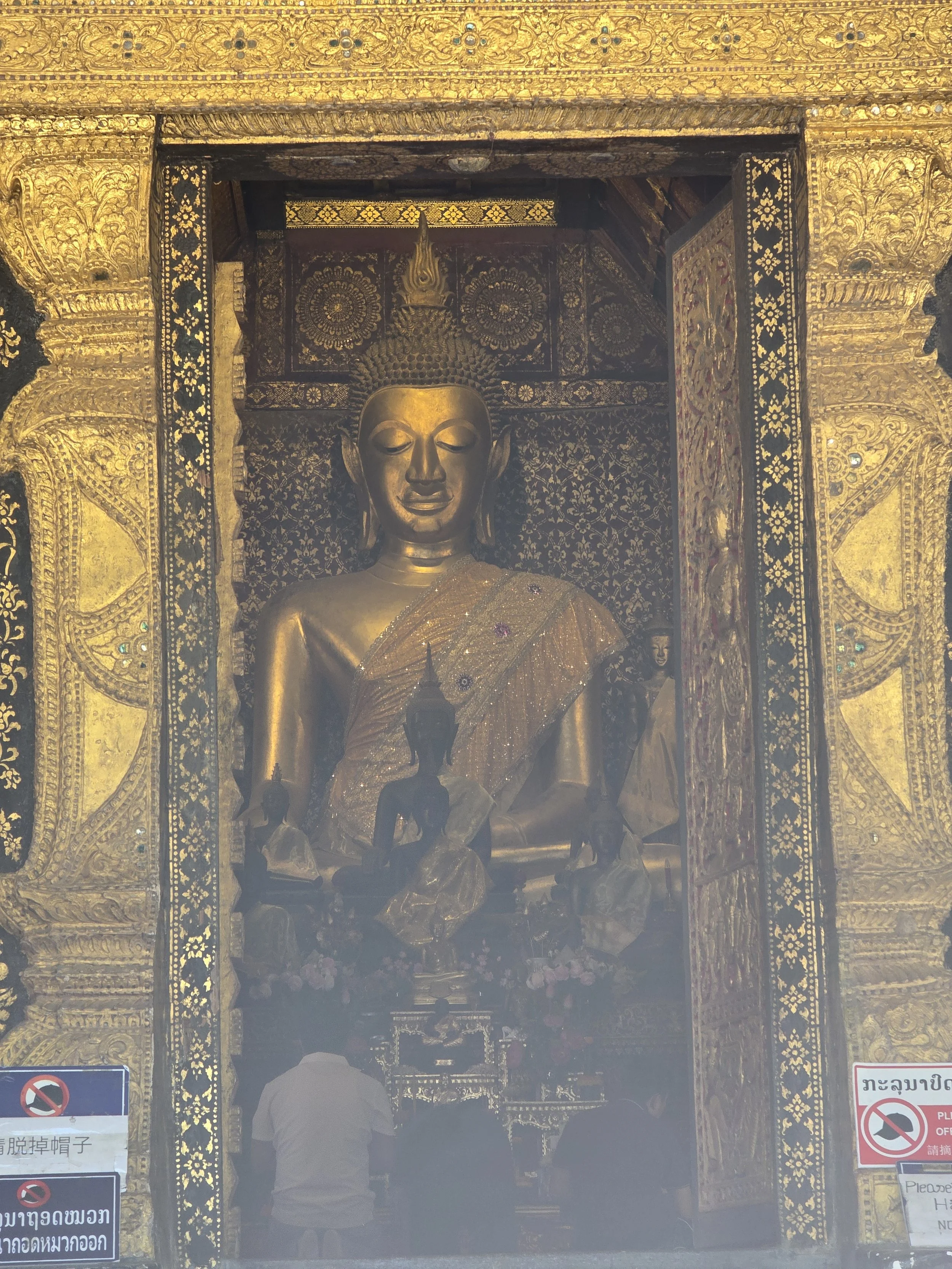 Statue dorée d'une figure religieuse dans un temple orné de motifs complexes en or. Deux visiteurs ou fidèles se trouvent devant la statue, certains portant des chapeaux.