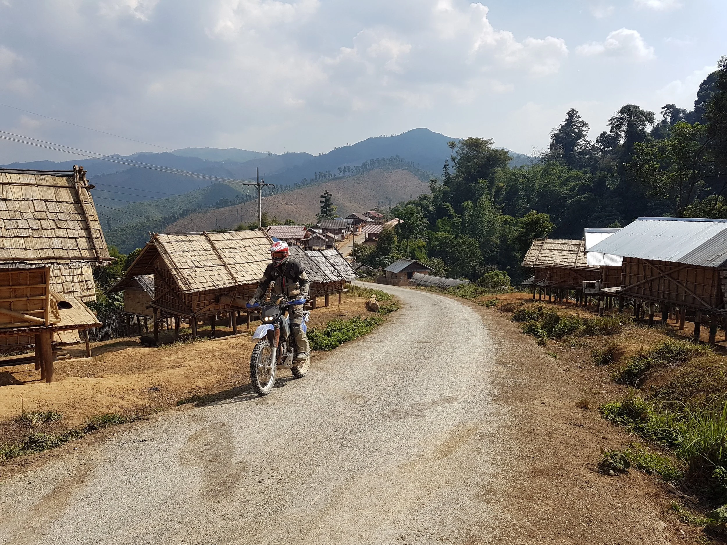 Vue d'un village rural avec maisons en bois, un chemin de terre, et des montagnes en arrière-plan, un homme portant un casque de moto. Scene en pleine journée avec ciel partiellement nuageux.