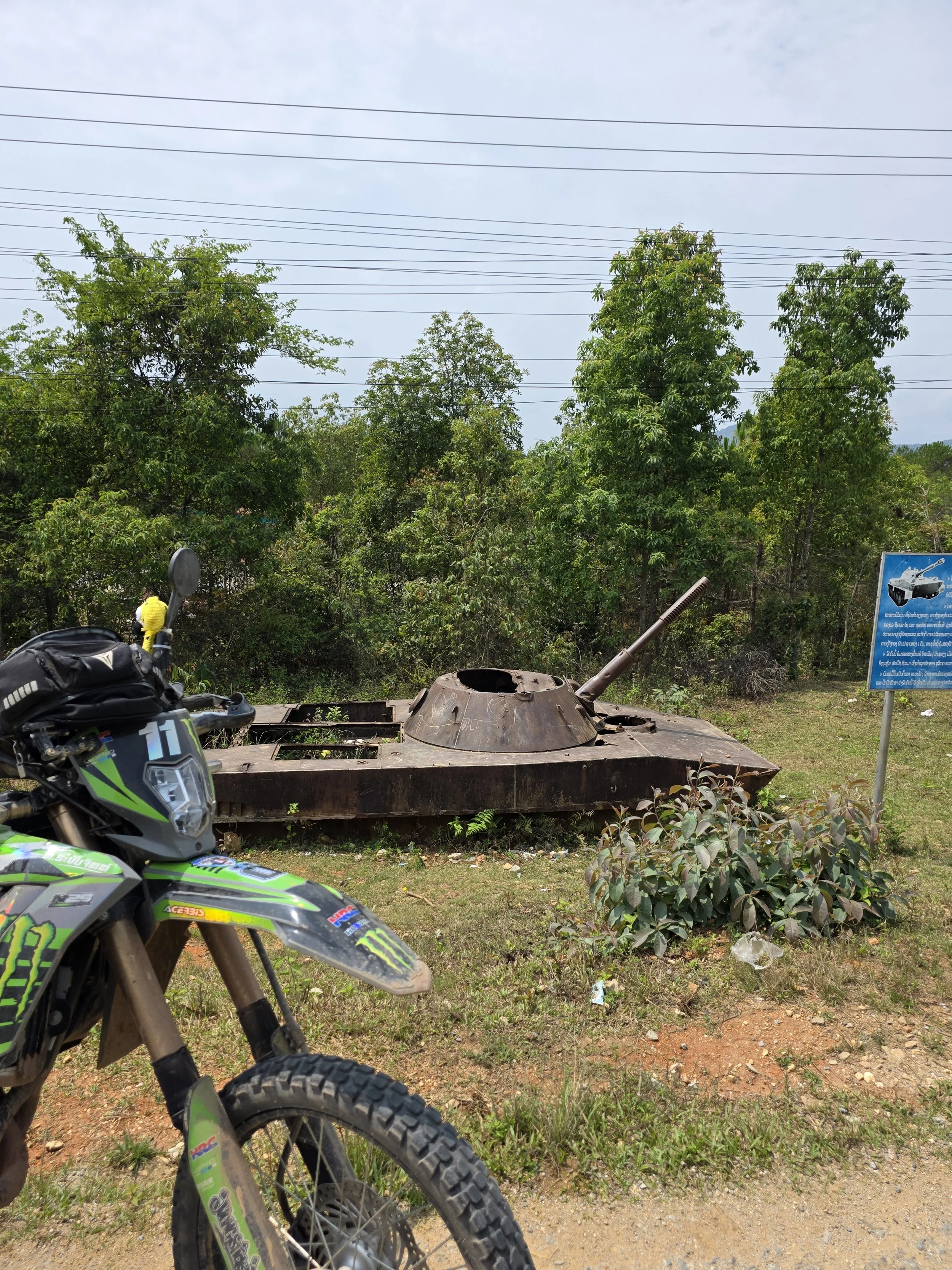 Une moto tout-terrain verte Garry avec un casque noir placé sur le guidon, un tank de char abandonné en métal rouillé, et un panneau d'information en arrière-plan, dans un environnement verdoyant avec des arbres et une route en terre.