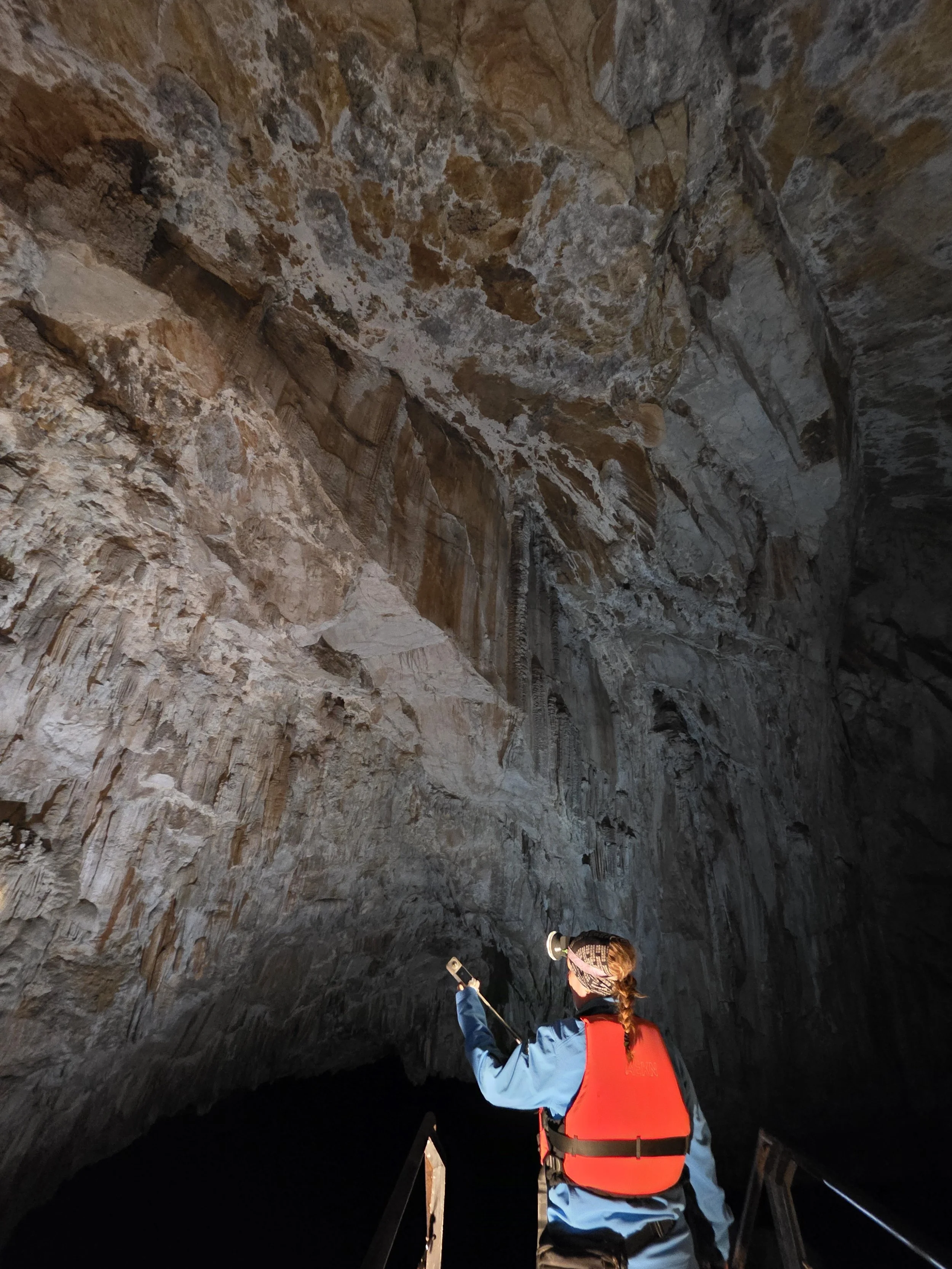 Une personne portant un chapeau, une veste bleue et un gilet de sauvetage orange explore une grotte ainsi qu'une autre personne, tenant une lampe torche. La grotte a des murs rocheux naturels.