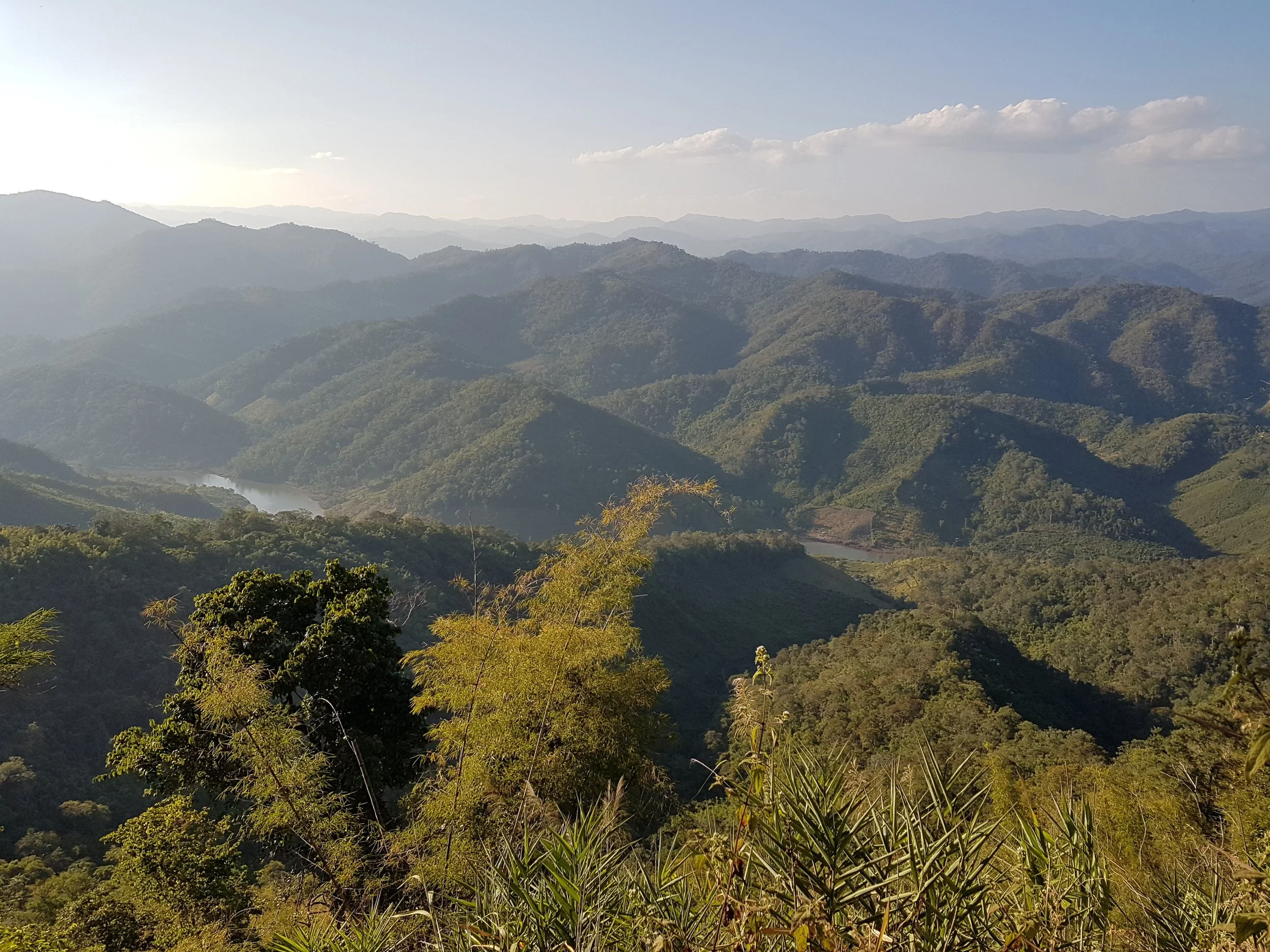 Paysage de montagnes verdoyantes avec un fleuve serpentant au fond, ciel clair avec quelques nuages.