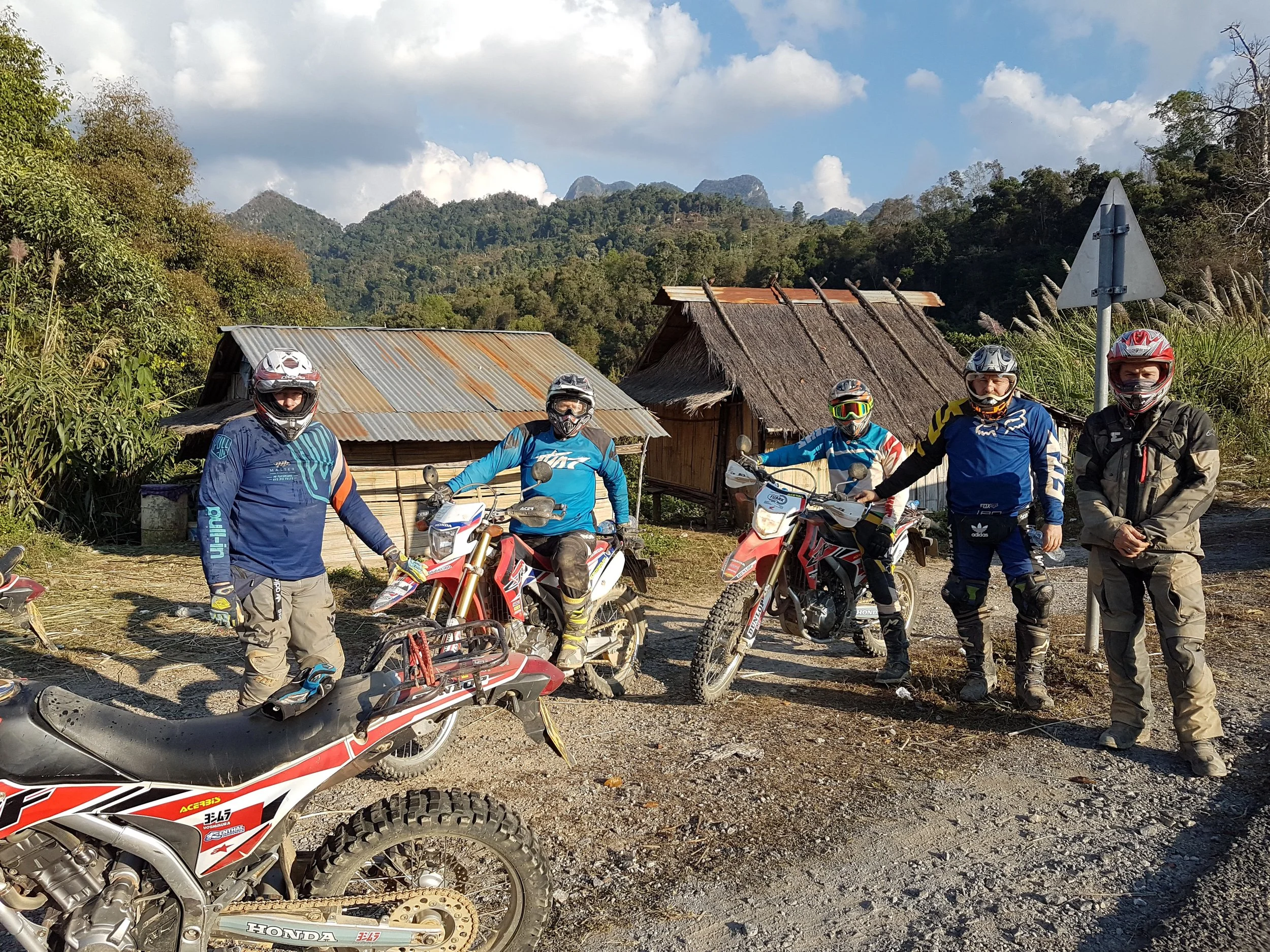 Groupe de cinq motocyclistes portant des équipements de protection, posant avec leurs motos tout-terrain dans un environnement rural entouré de montagnes et de maisons en bois avec un ciel partiellement nuageux.
