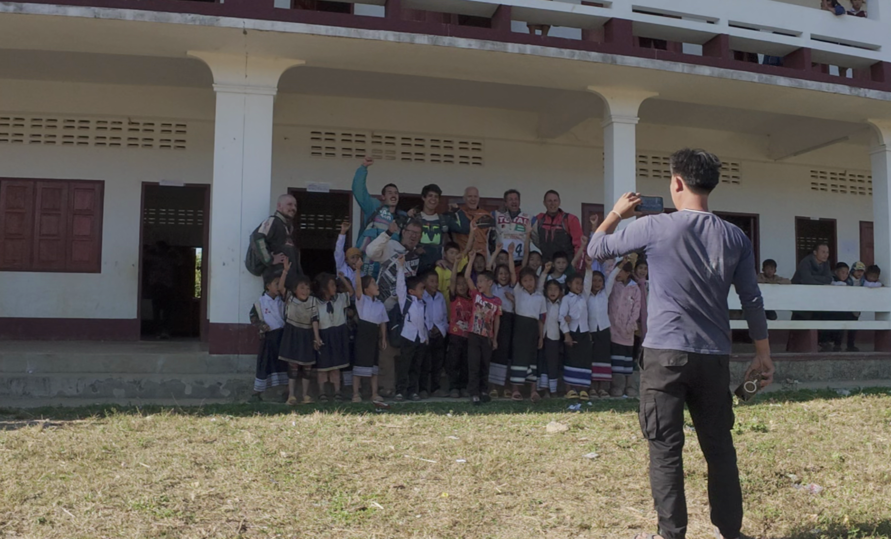Un groupe d'enfants et d'adultes pose pour une photo devant un bâtiment scolaire. Un homme prend la photo à distance, et un groupe de personnes se trouve à l'étage.