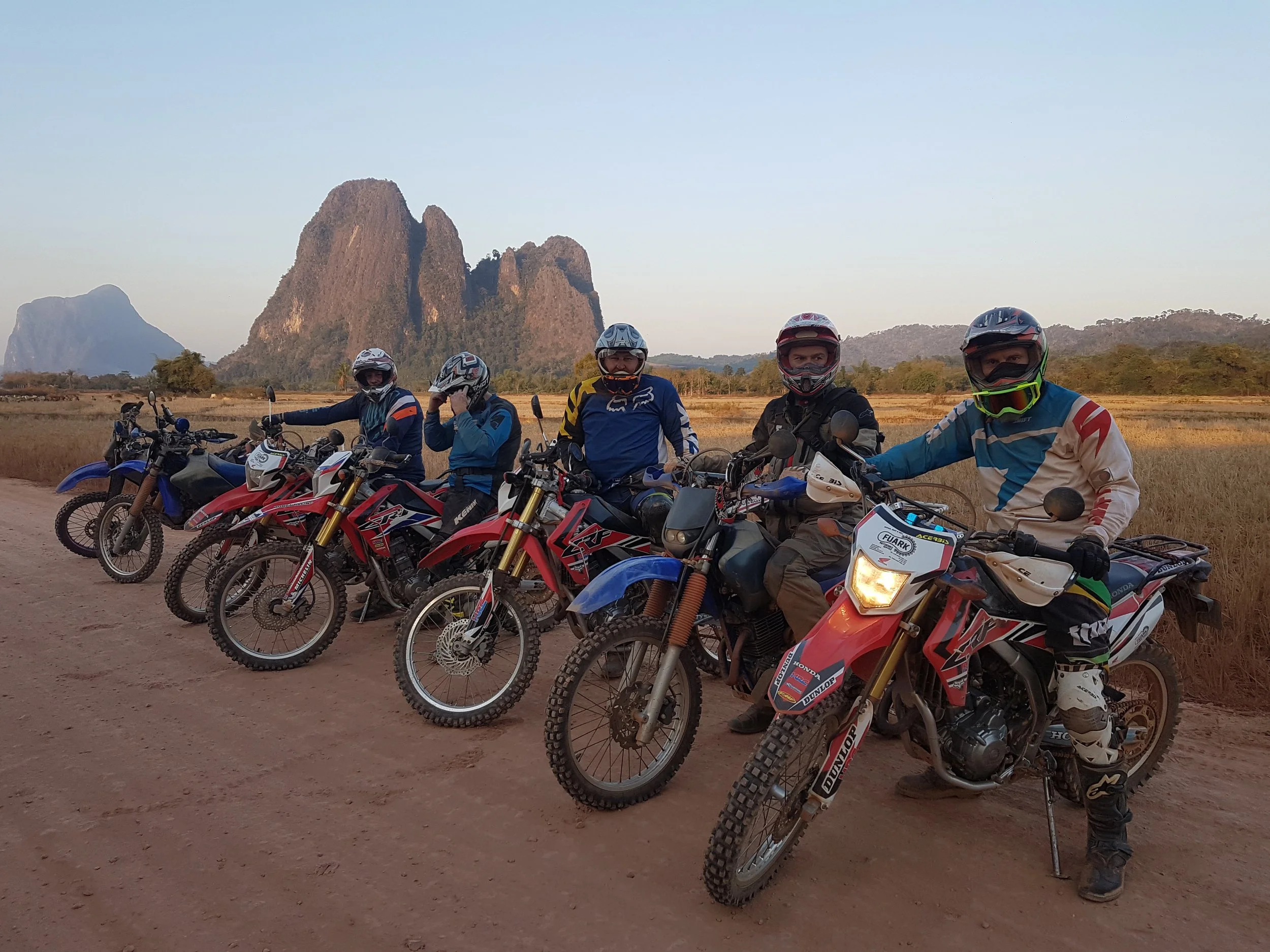 Groupe de cinq motards en tenue de motocross, portant des casques, débout devant leurs motos tout-terrain, avec un paysage de champs et des montagnes en arrière-plan, sous un ciel clair.