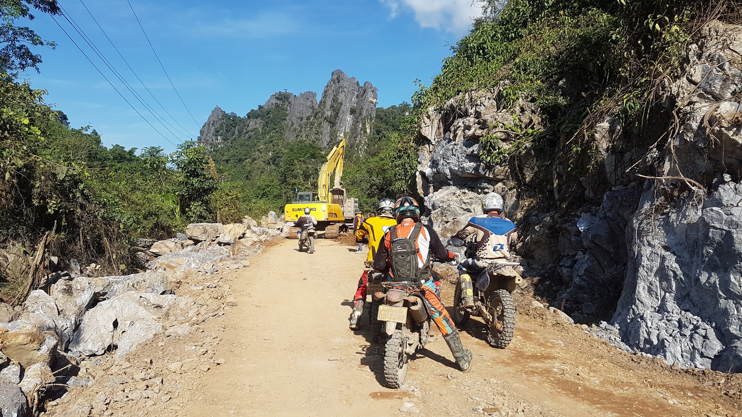 Groupe de motards en plein voyage sur une route de montagne, entourés de rochers et de végétation verdoyante, avec une grande formation rocheuse visible à l'arrière-plan.