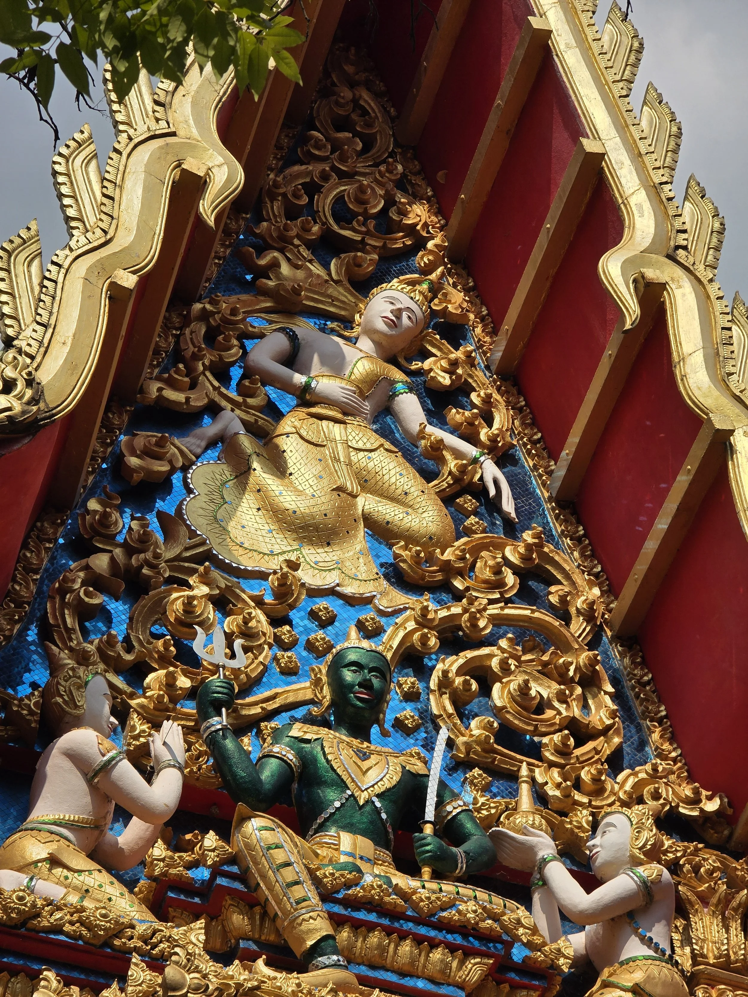 Une sculpture ornée de dorure représentant une figure humaine dans un style traditionnel, avec d'autres figures en dessous. La sculpture est richement décorée avec des motifs complexes et encadrée par un cadre doré et rouge.