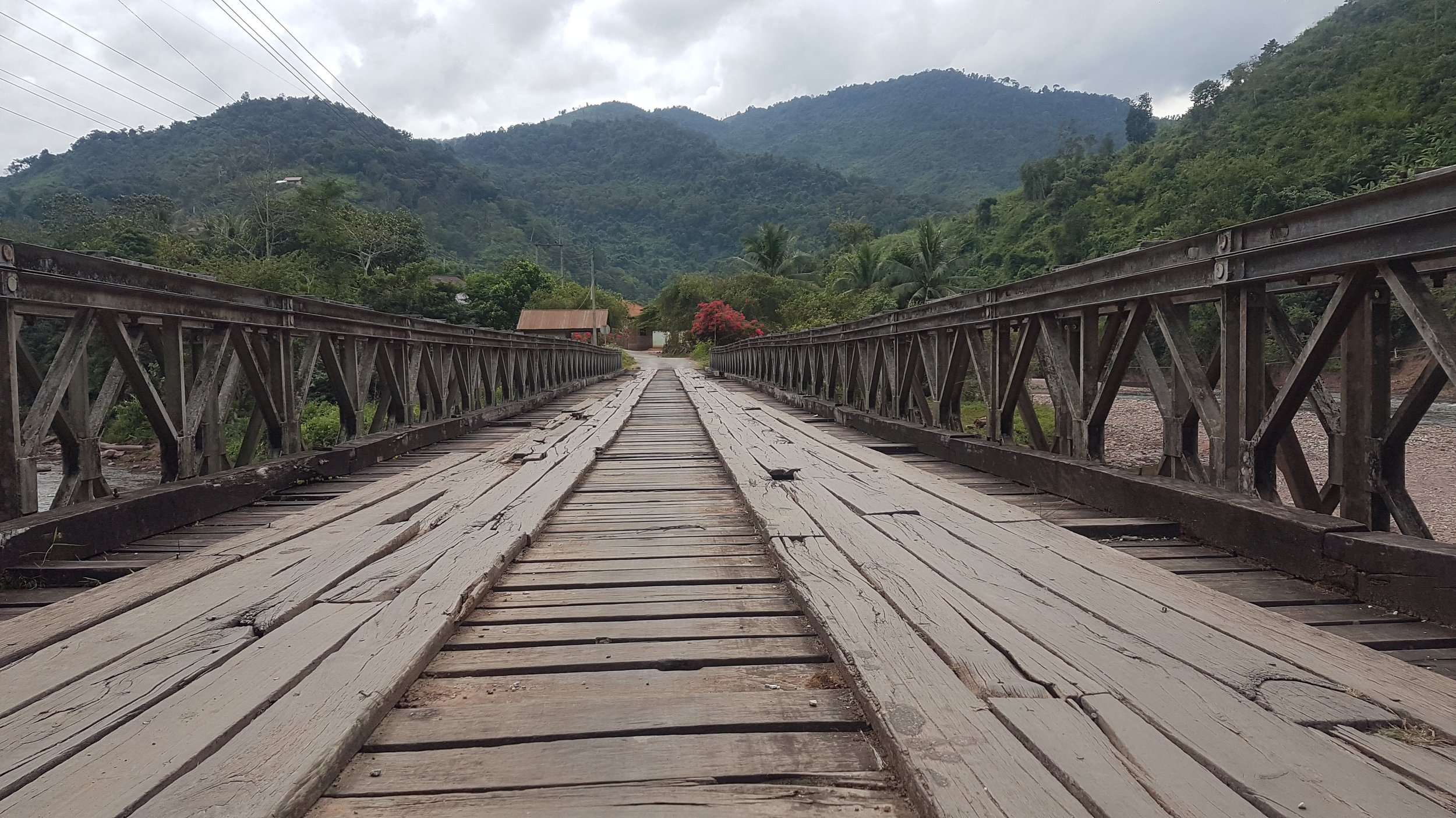 Un pont en bois vieux, traversant une rivière, avec des montagnes verdoyantes à l'arrière-plan et un ciel nuageux.
