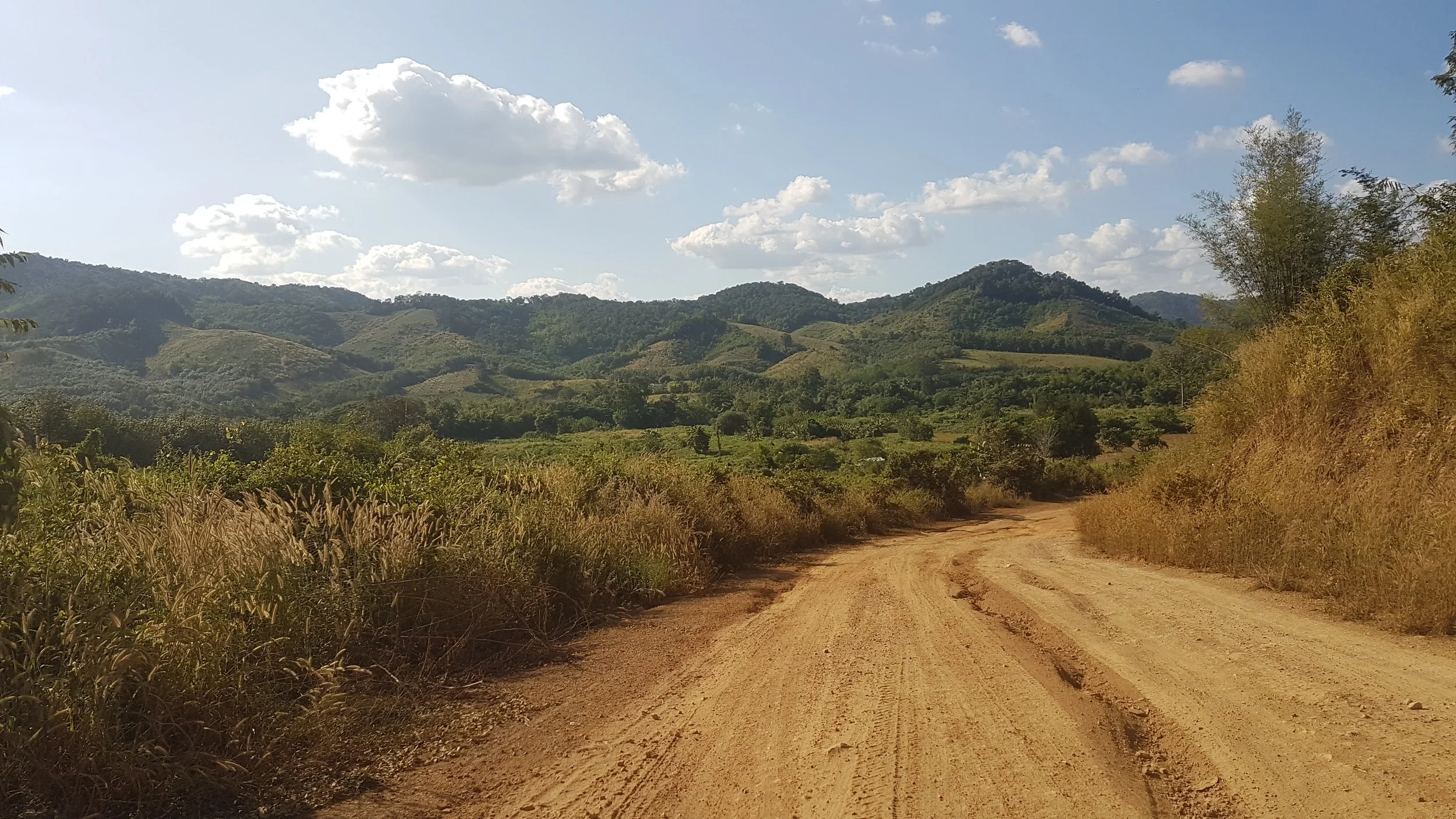 Une route en terre serpentant à travers un paysage de collines verdoyantes sous un ciel partiellement nuageux.